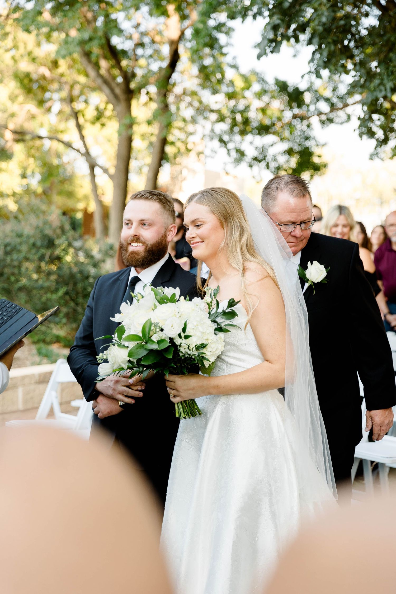 Wedding party members walking in a line outdoors carrying white bouquets during a ceremony.