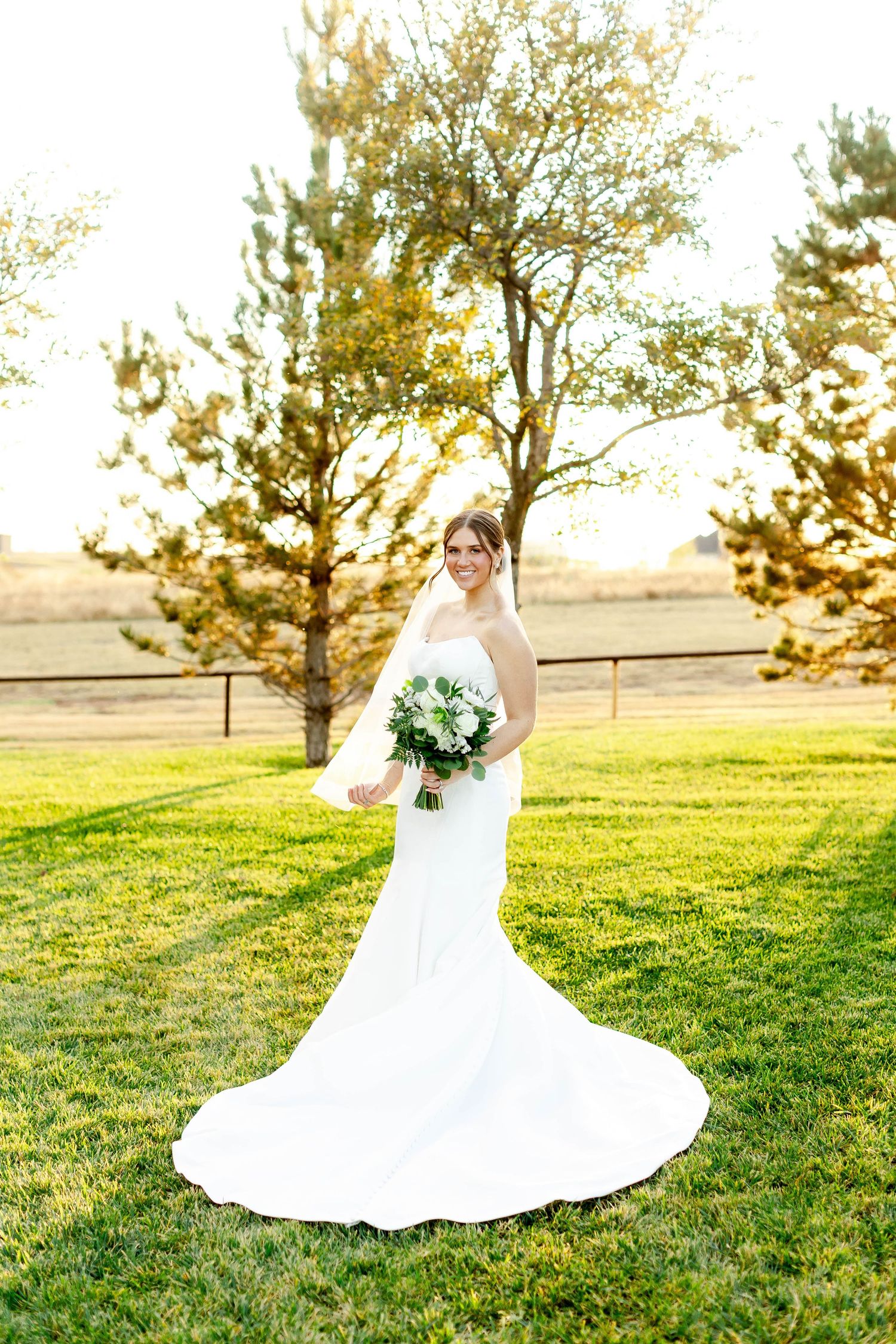 A bride in a flowing white dress poses in a grassy field at golden hour.
