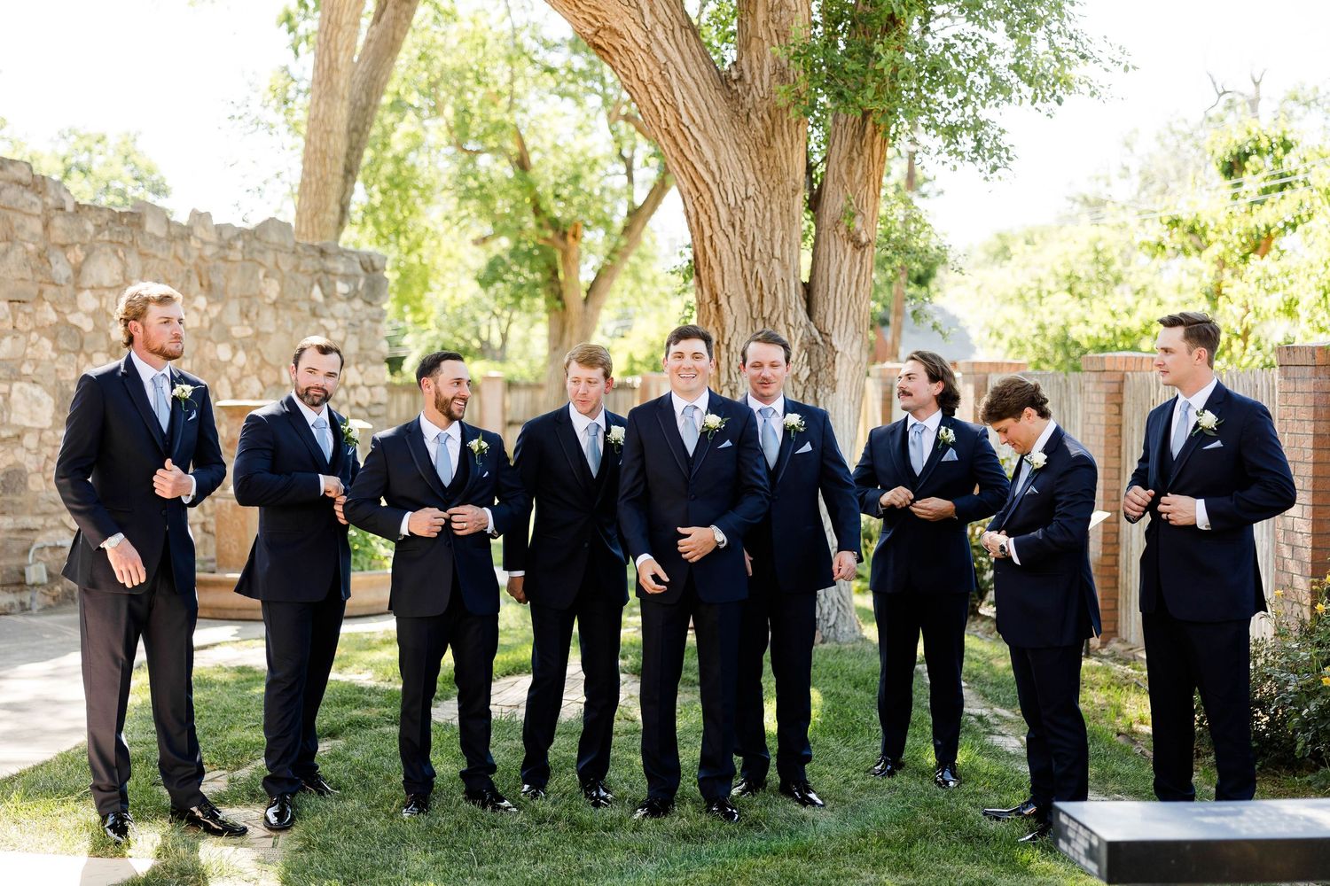 Groomsmen in dark suits posing casually outdoors under trees.