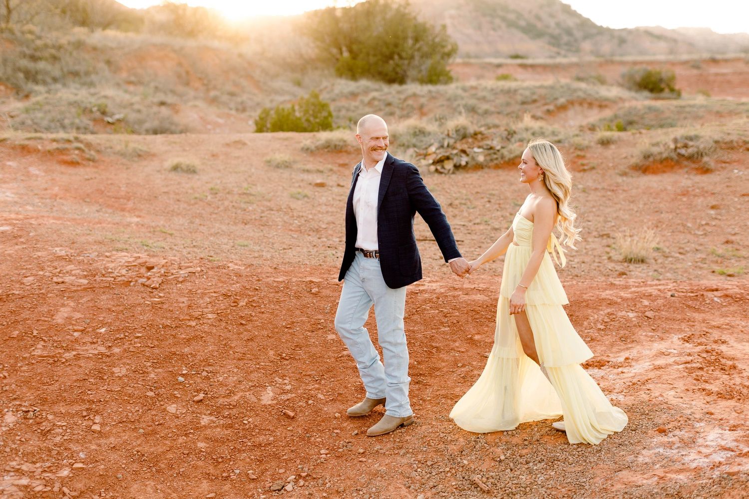 A couple walks hand in hand across red desert terrain during golden hour.