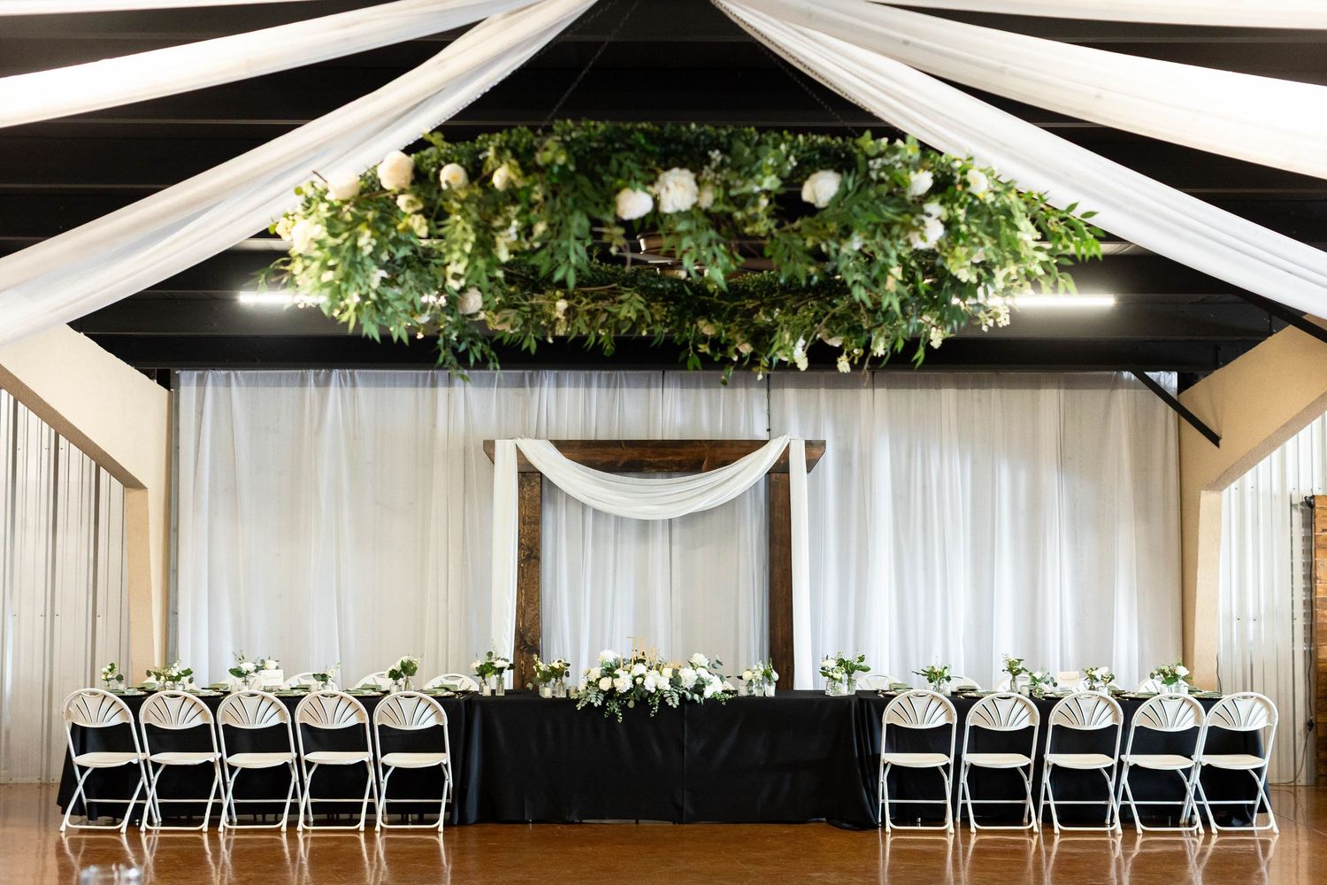 Black and white chairs line up in rows beneath elegant white draping and greenery at an indoor wedding venue.