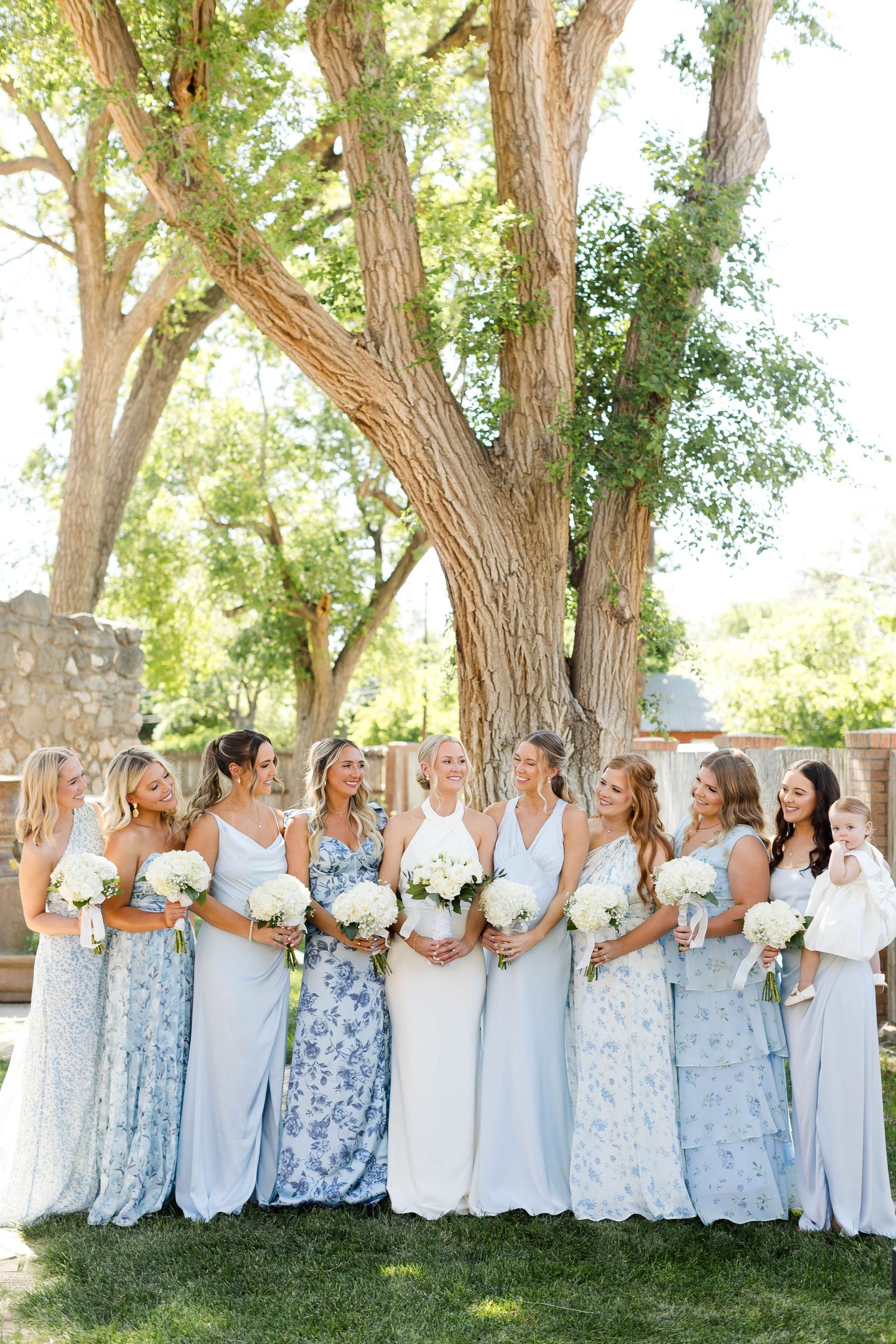 Large bridal party in coordinating blue and white dresses posed under trees.