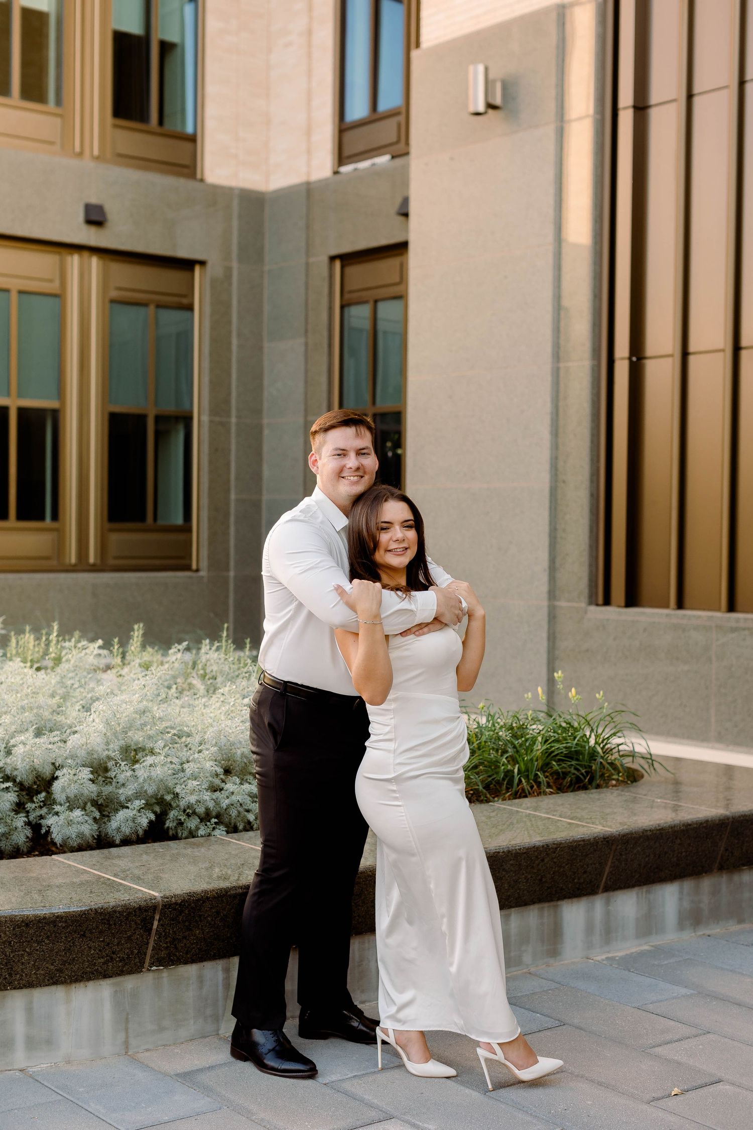 Couple in formal attire embracing outside modern building with white dress and dress shirt.