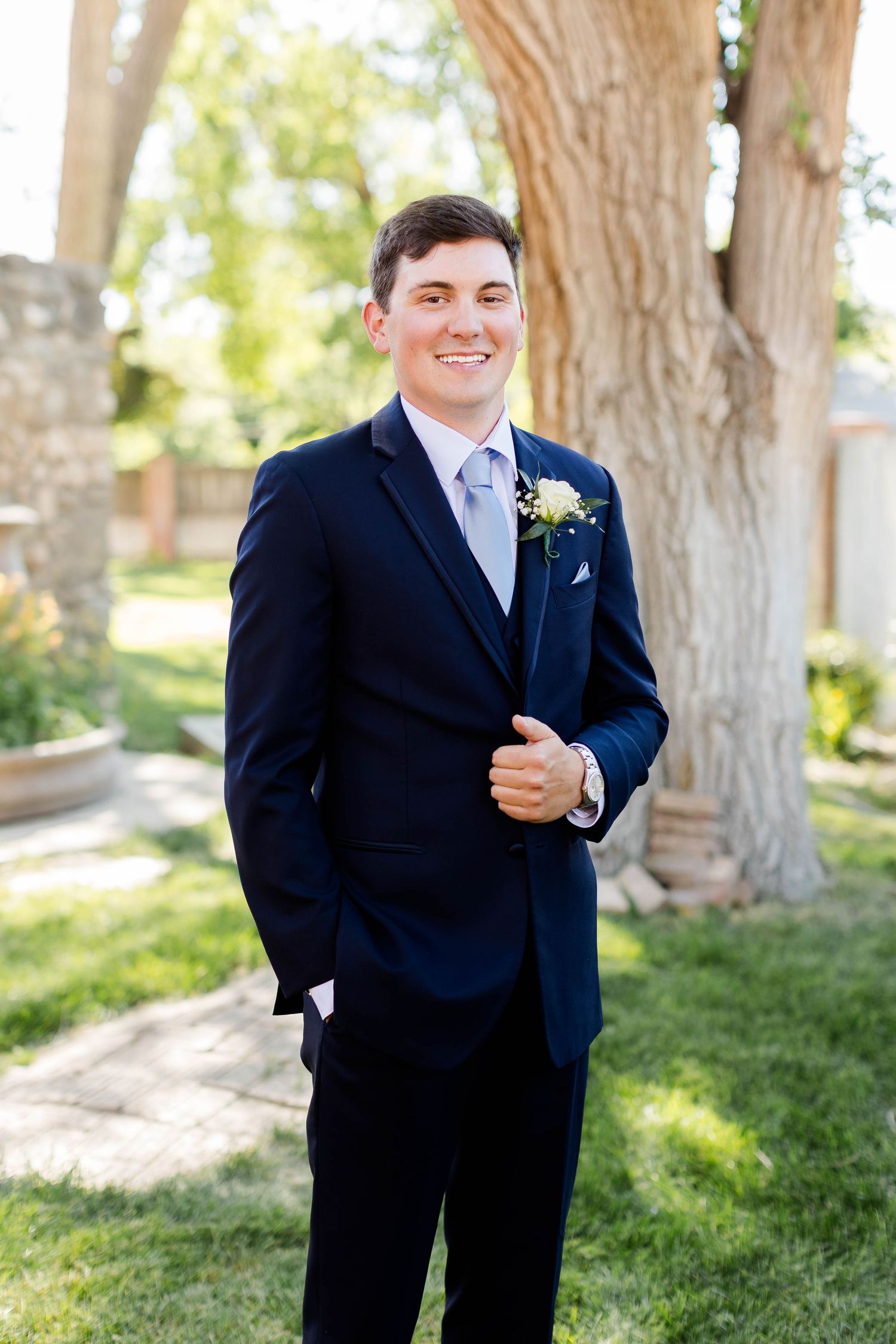A person in a navy suit with boutonniere poses outdoors on grass near trees.