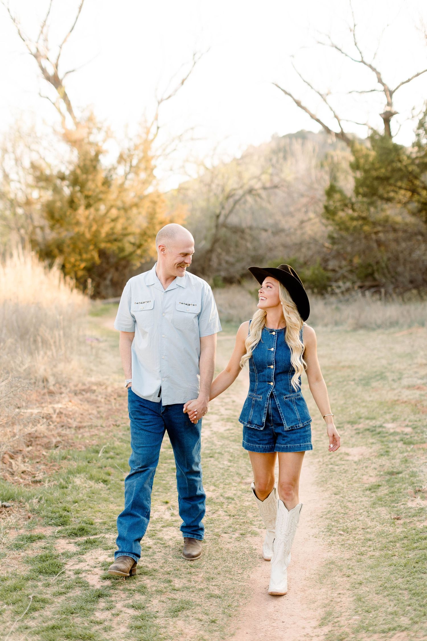 Couple in denim and cowboy boots walk hand in hand on a desert path at sunset.