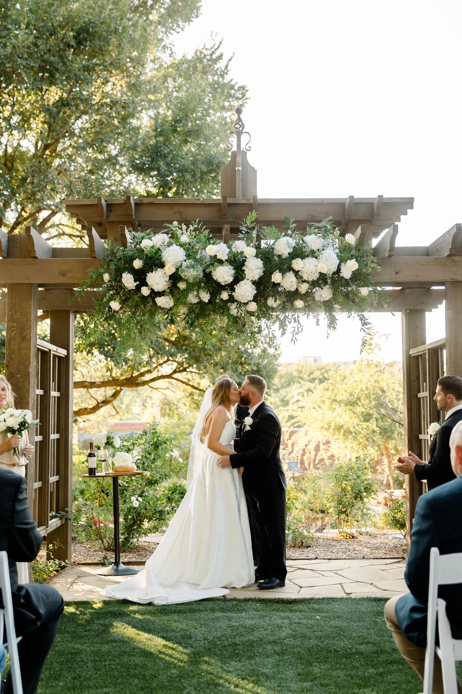 Scenic wedding ceremony setup with wooden pergola and white floral arrangements against natural landscape.