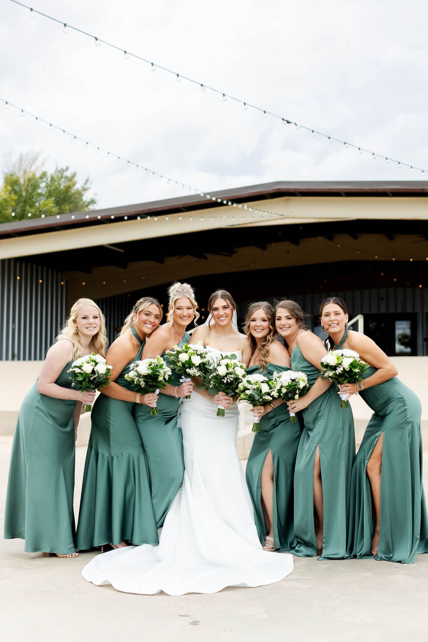 Wedding party wearing sage green dresses and white bridal gown share laughter during outdoor photo session.