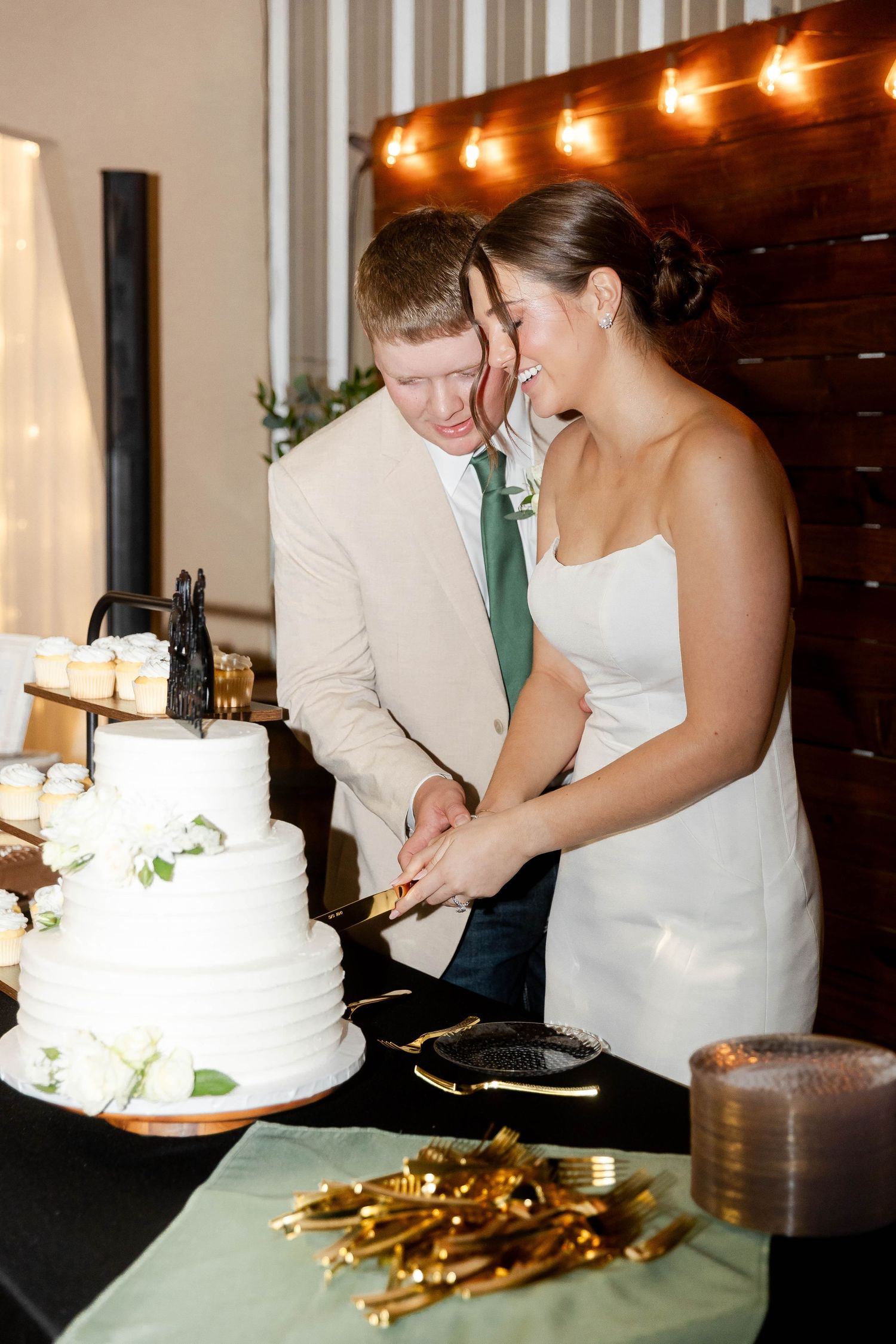 A couple cuts their white wedding cake together under warm indoor lighting.