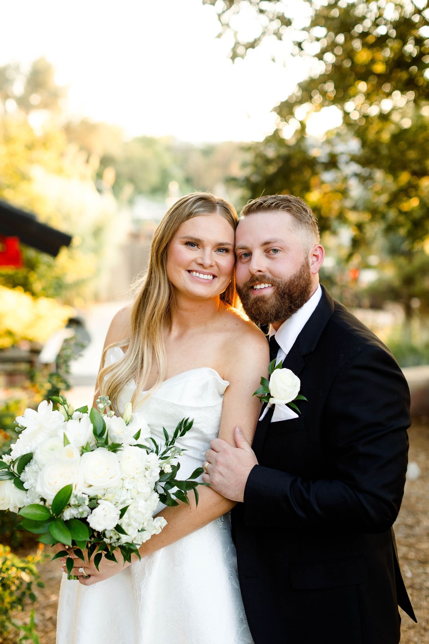 A couple in wedding attire pose together outdoors with a bouquet of white flowers on their special day.