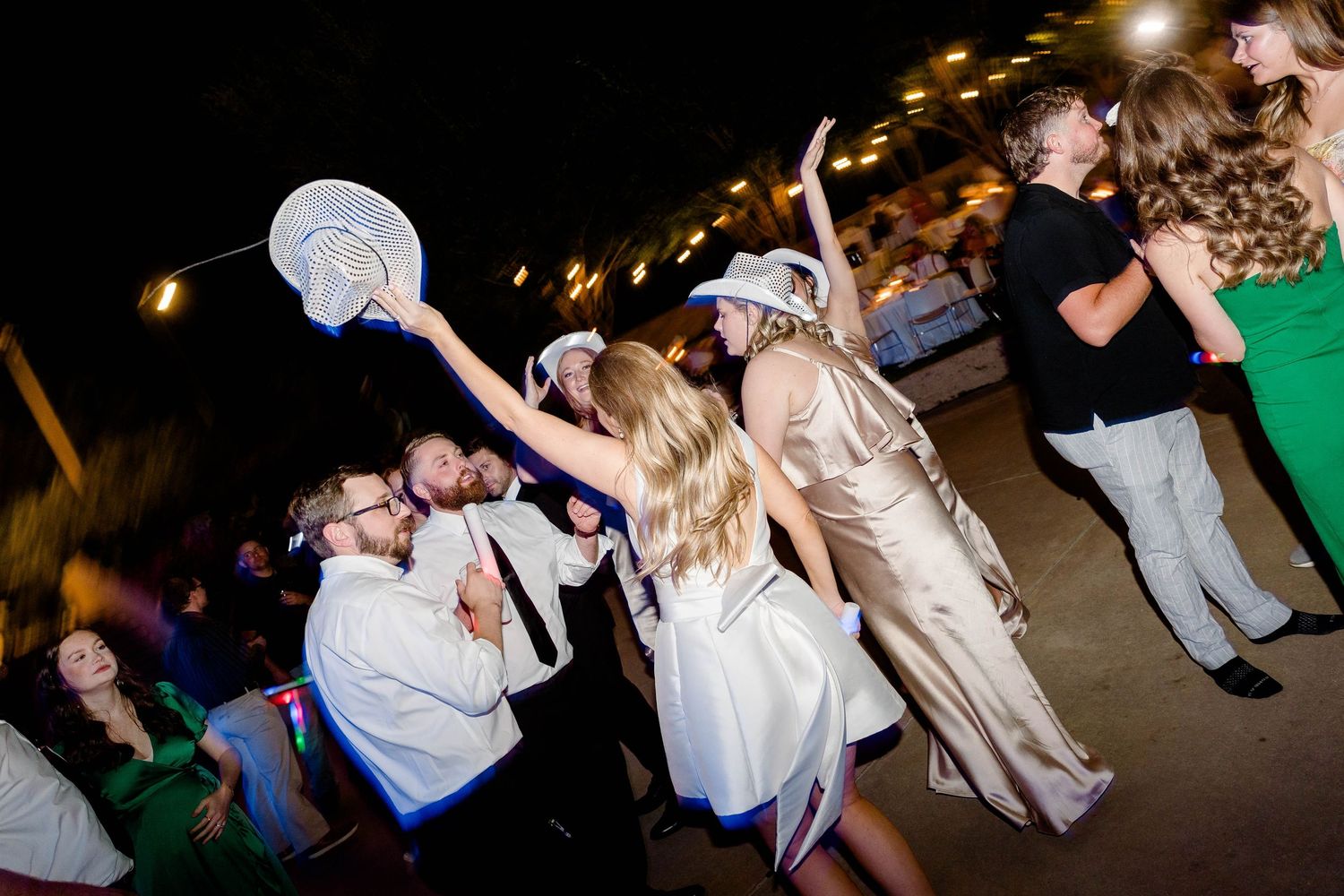 Wedding party dancing at night under decorative string lights at an outdoor reception.