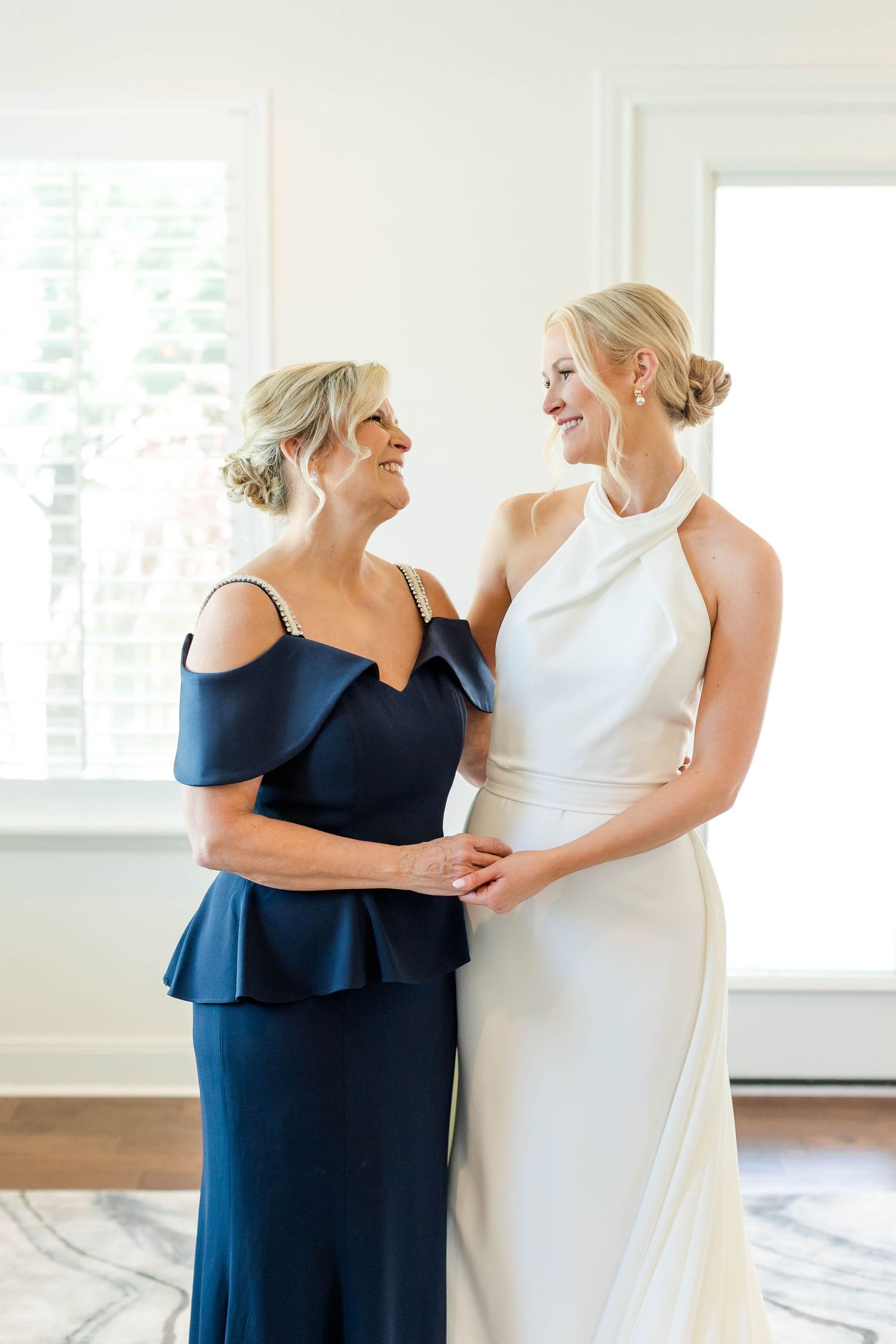 Two women share an emotional moment in formal dresses during wedding preparations.
