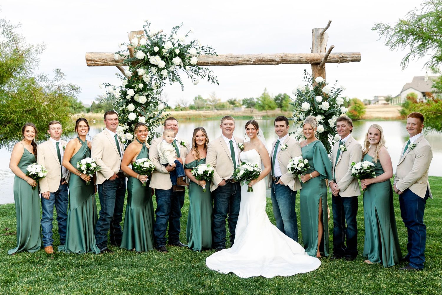 Wedding party poses in sage green and beige attire under a rustic wooden arch with white floral decorations.