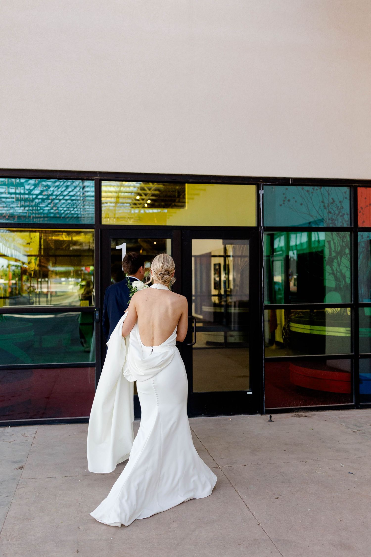 A bride in a flowing white dress walks outside a modern building with glass windows.