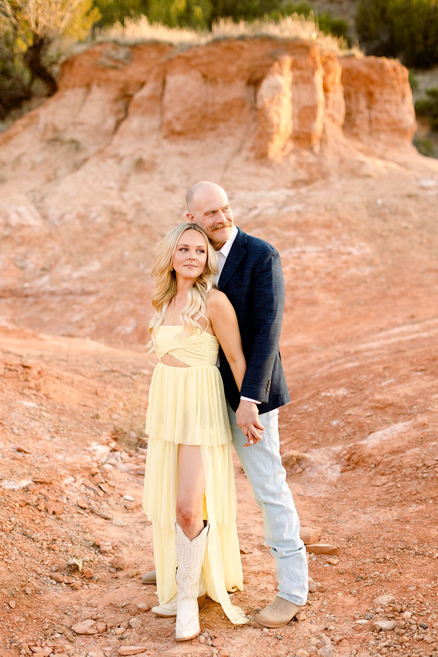 A couple poses together in western attire against red rock formations during sunset.
