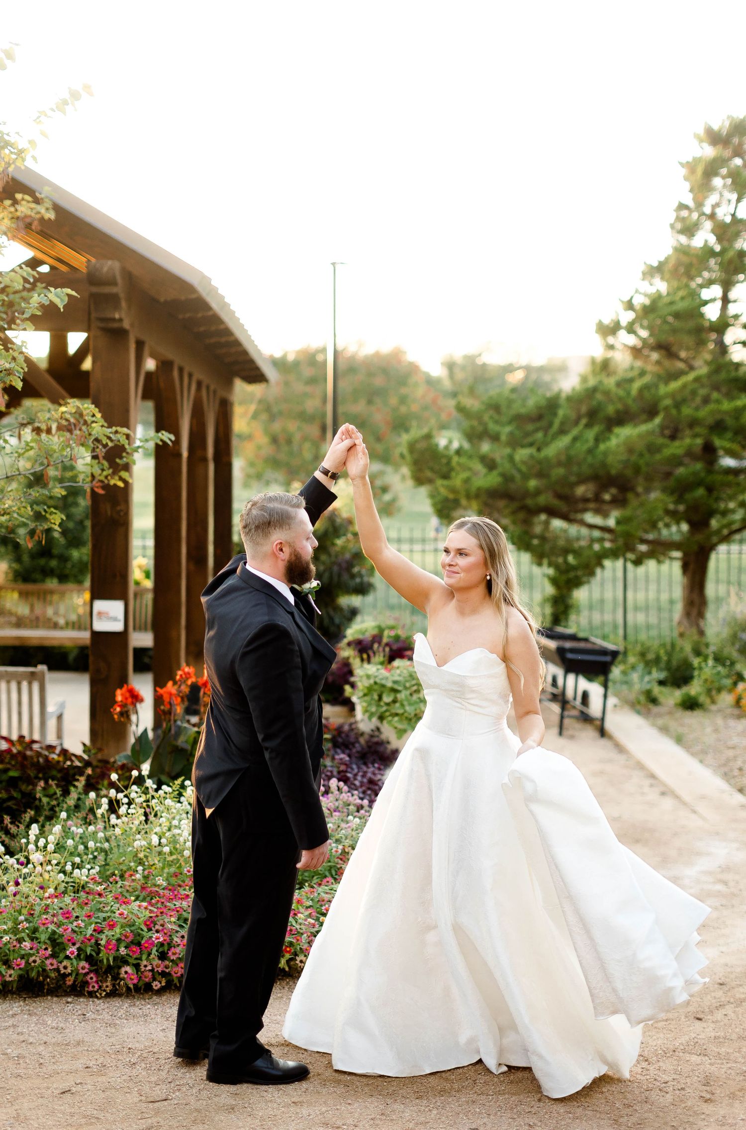 Wedding couple dances playfully under wooden pergola surrounded by garden flowers.
