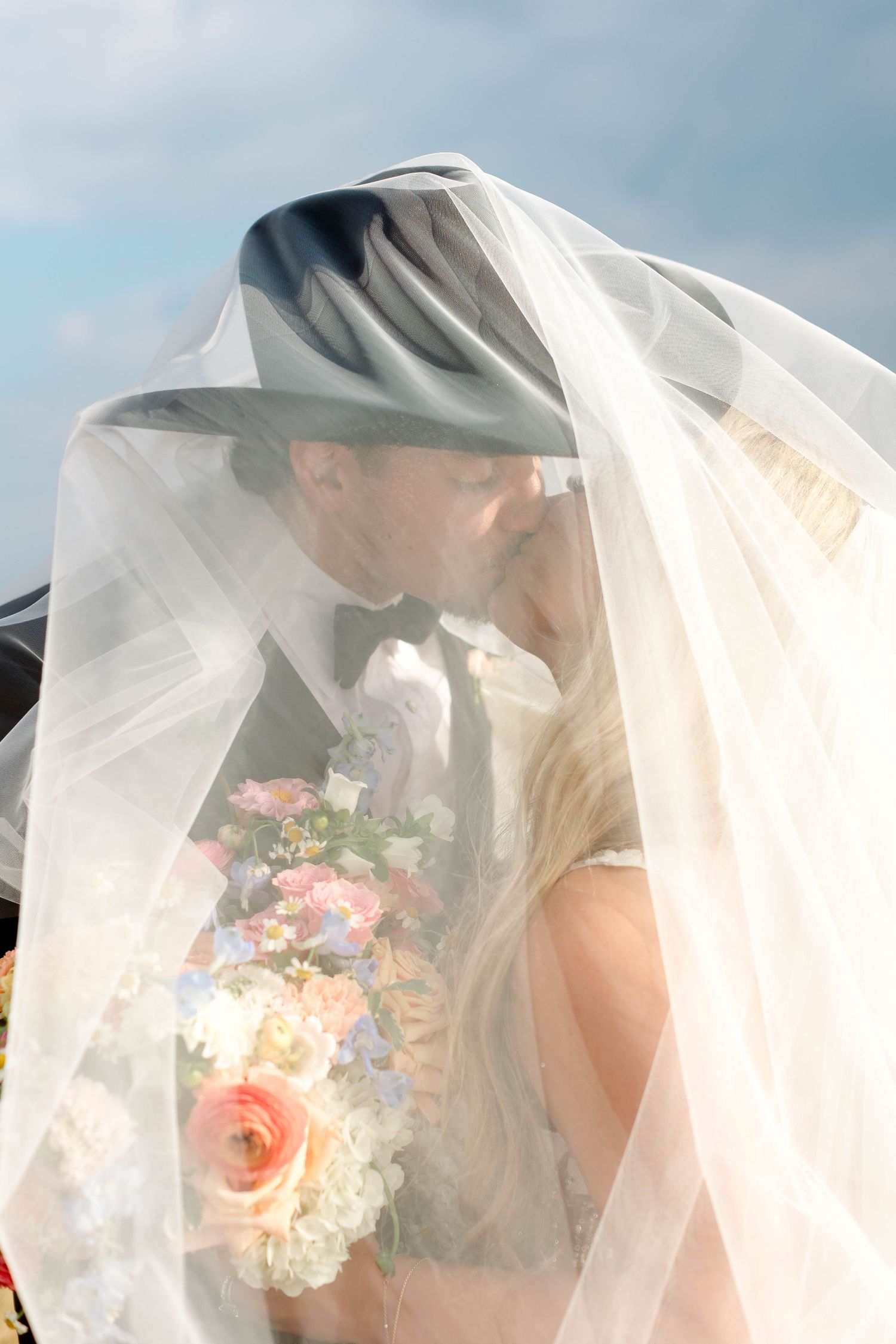 Ethereal bridal portrait with flowing veil and colorful bouquet against blue sky.