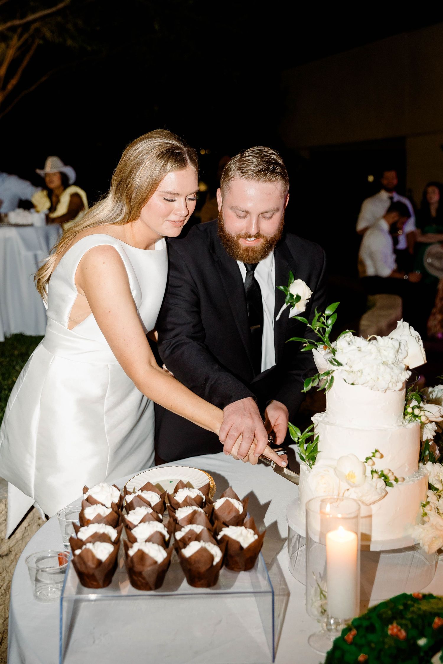 Couple cutting their elegant white wedding cake with chocolate desserts displayed on the table.