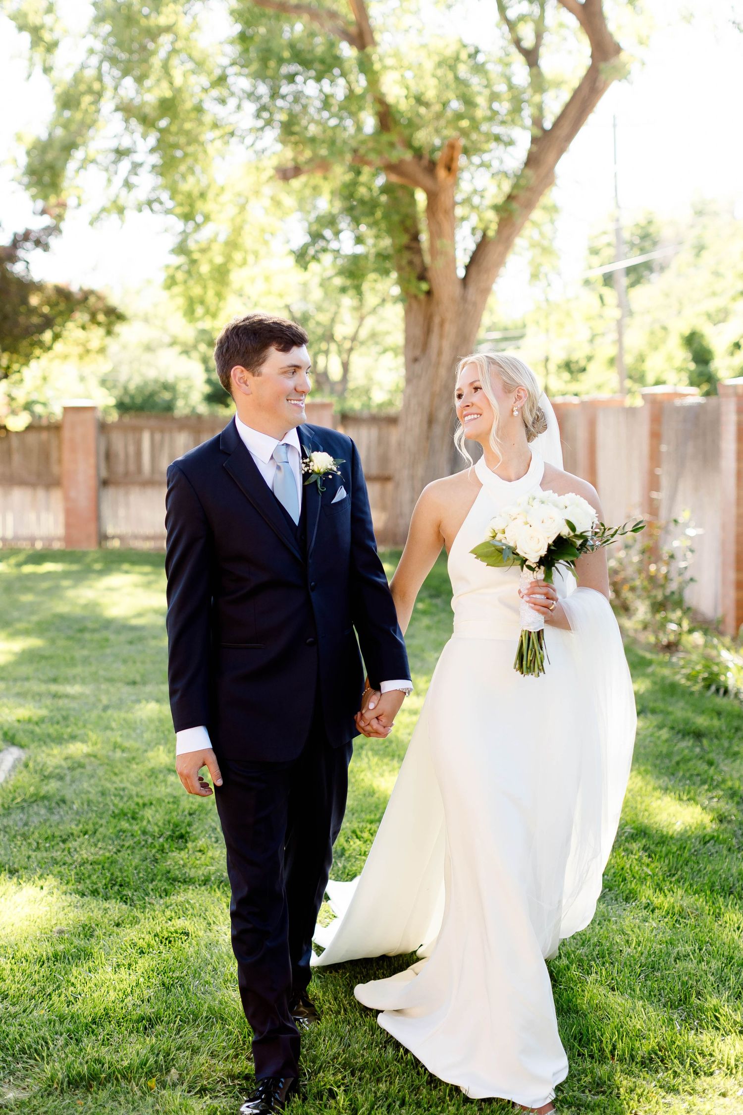 Wedding couple strolling through a sunlit garden with beautiful trees and a wooden fence.