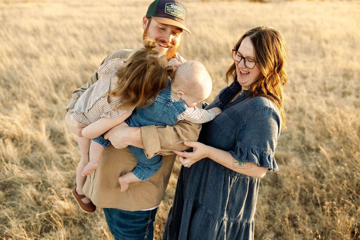 A couple in casual clothing walks together through a golden field during sunset holding their baby.