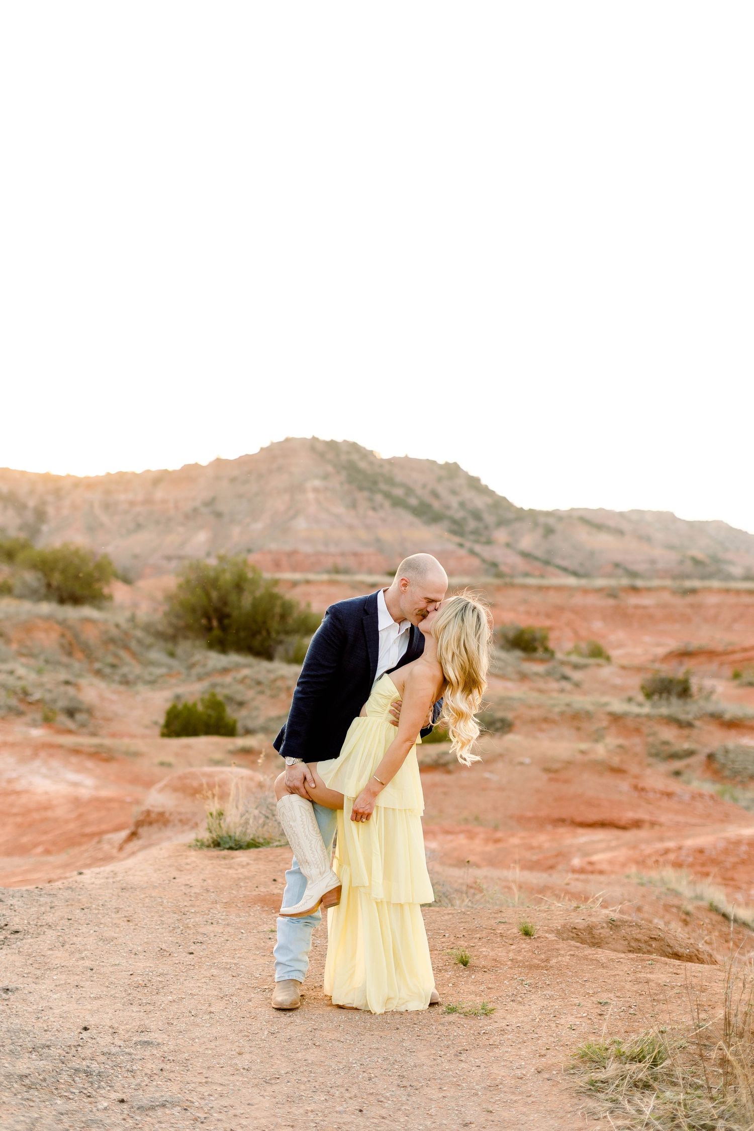 A couple in yellow dress and casual attire poses romantically against a desert landscape at sunset.