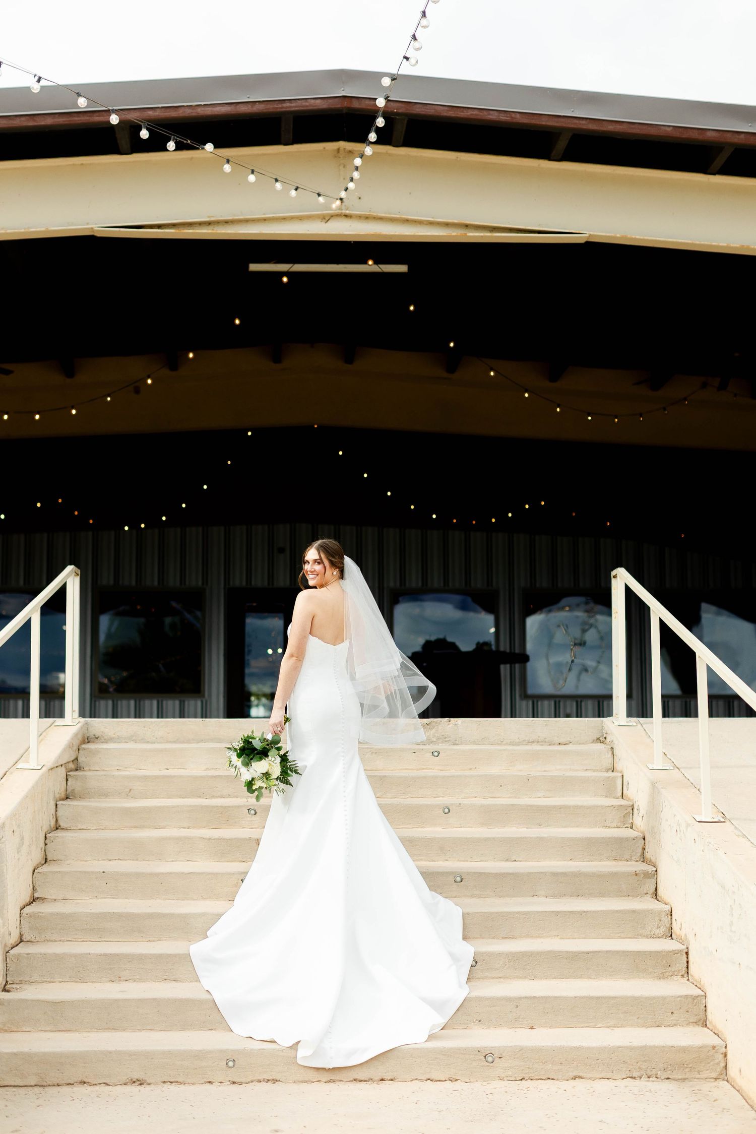 Wedding dress train flows elegantly across concrete steps with twinkle lights decorating the venue above.