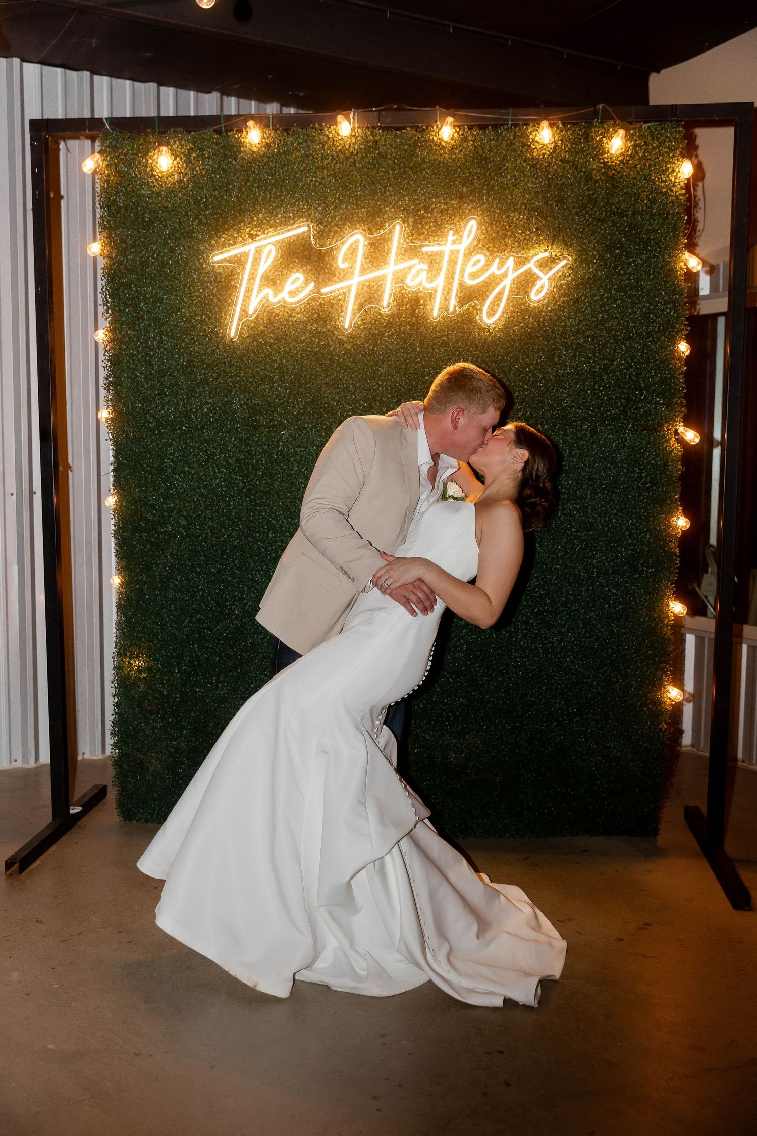 A couple performs a romantic dip kiss in front of an illuminated neon sign.