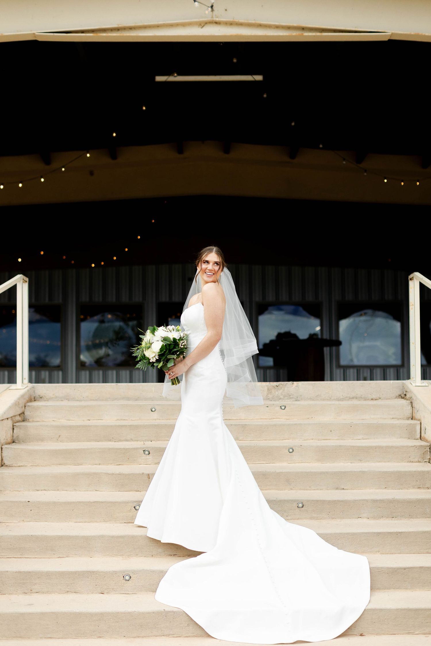A bride in a flowing white wedding dress poses gracefully on concrete steps with string lights in the background.
