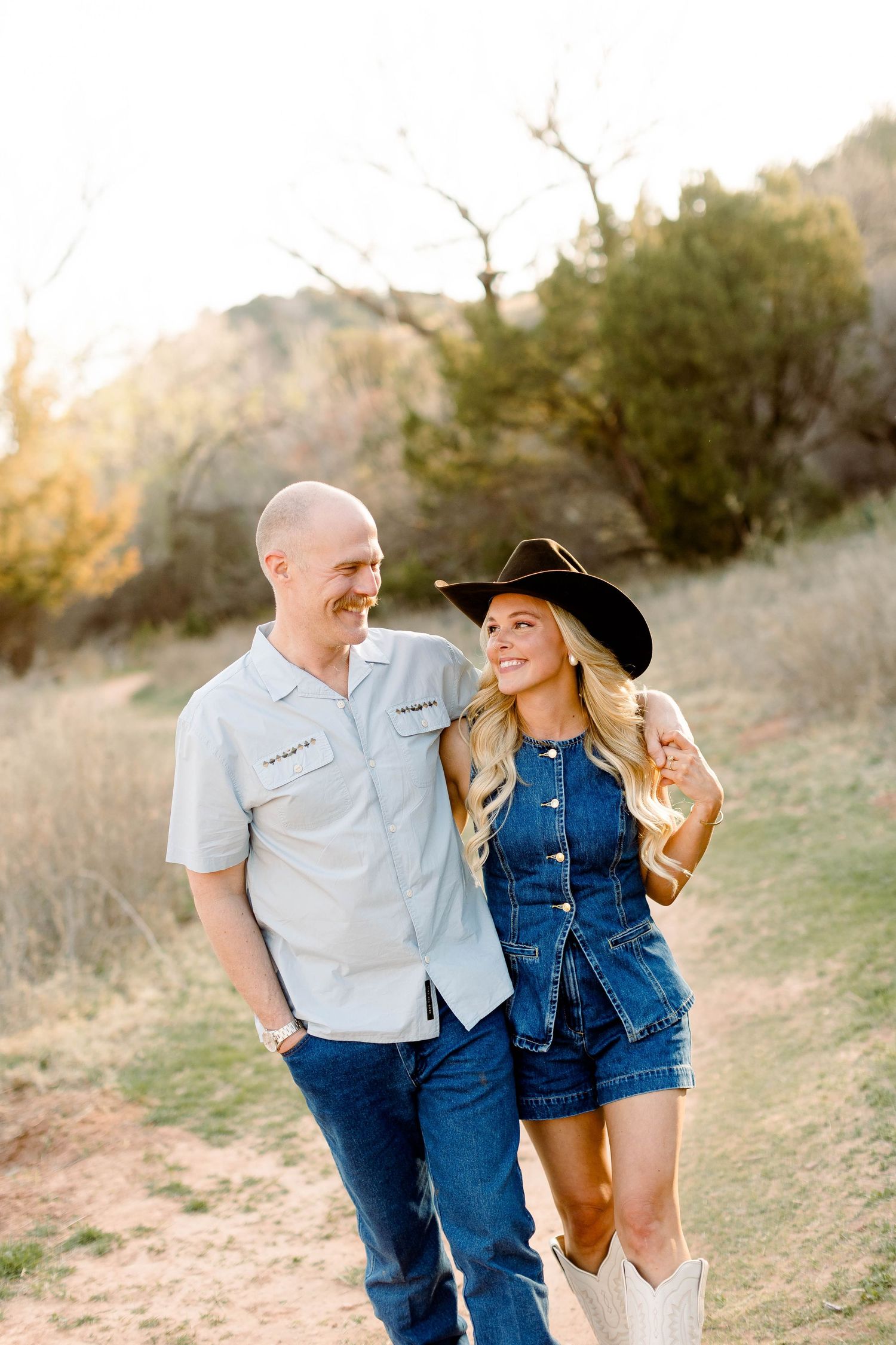 Couple in western-inspired outfits share laughs during sunset photoshoot on desert trail.