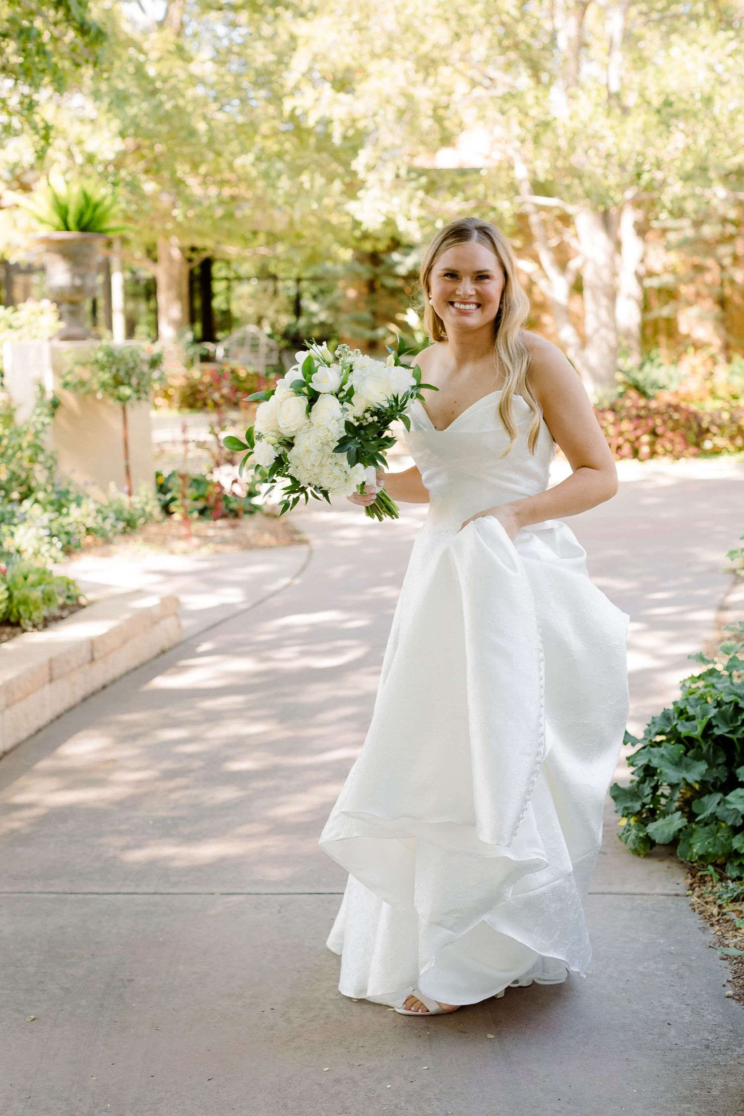 A bride in a flowing white wedding dress twirls playfully on a sunlit path surrounded by greenery.
