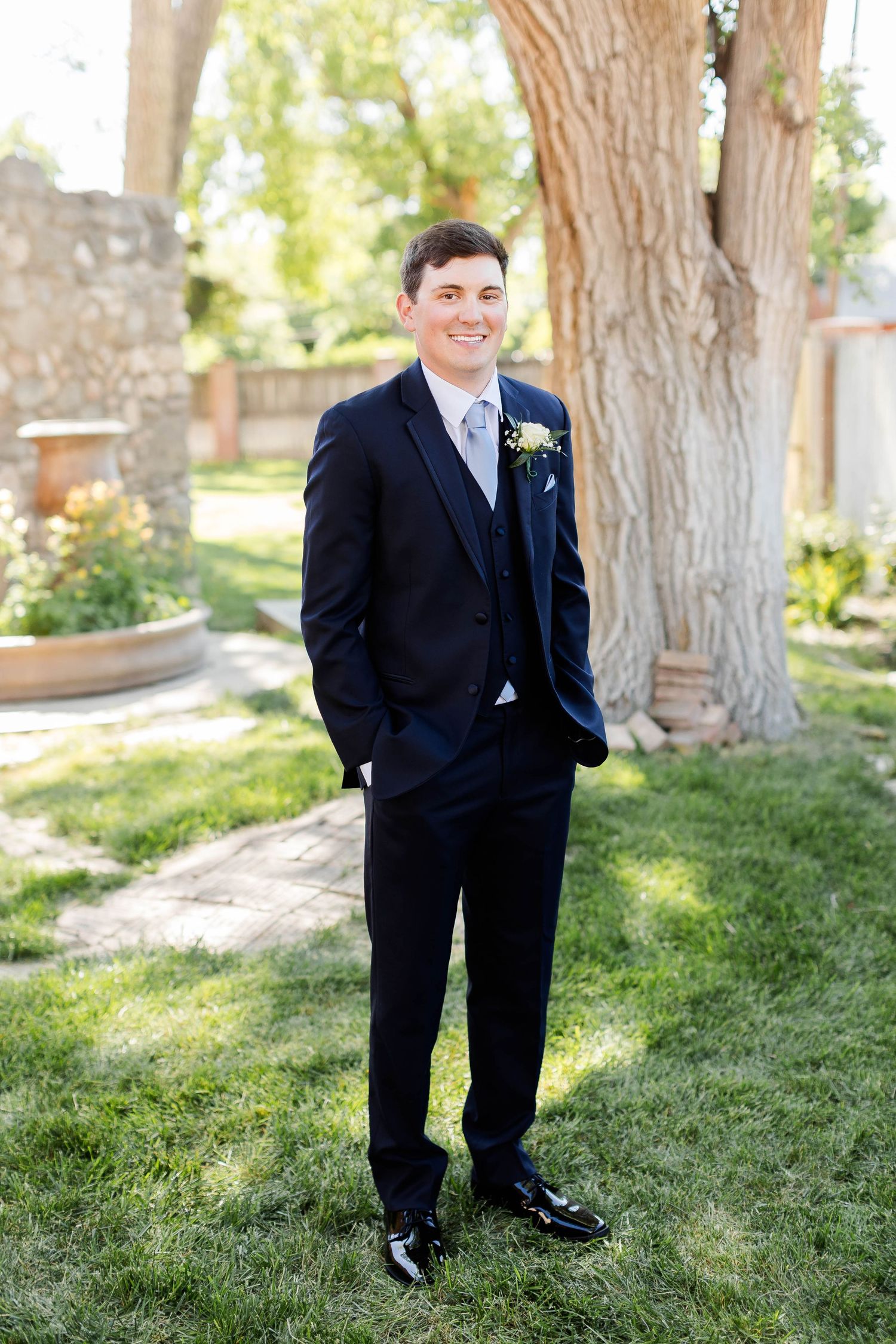 A person in a formal navy suit stands outdoors near a large tree.