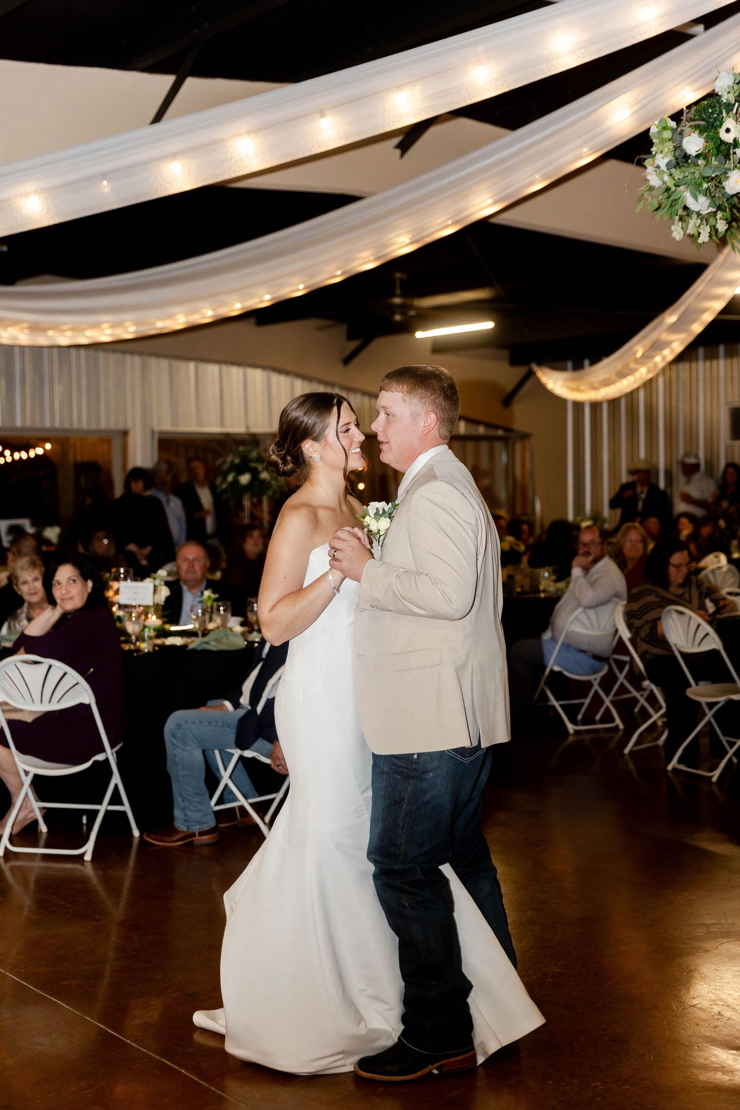 Wedding reception dance scene in a rustic barn venue decorated with white drapes and string lights.