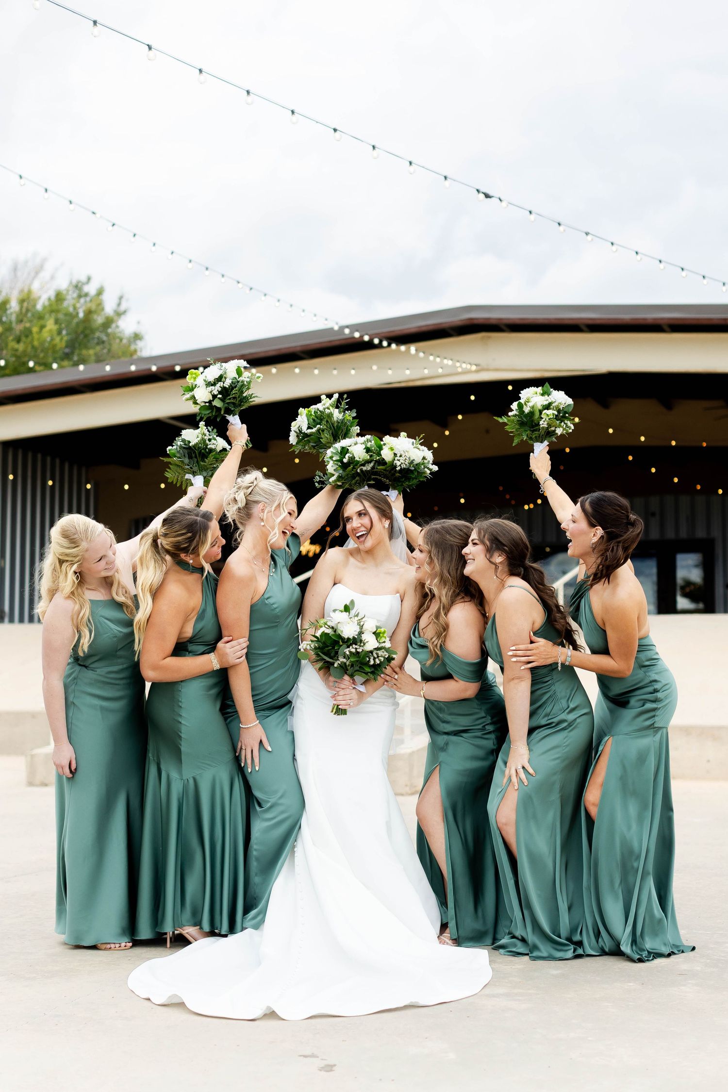 A bridal party in sage green bridesmaid dresses and white wedding gown pose together in a candid moment outdoors.