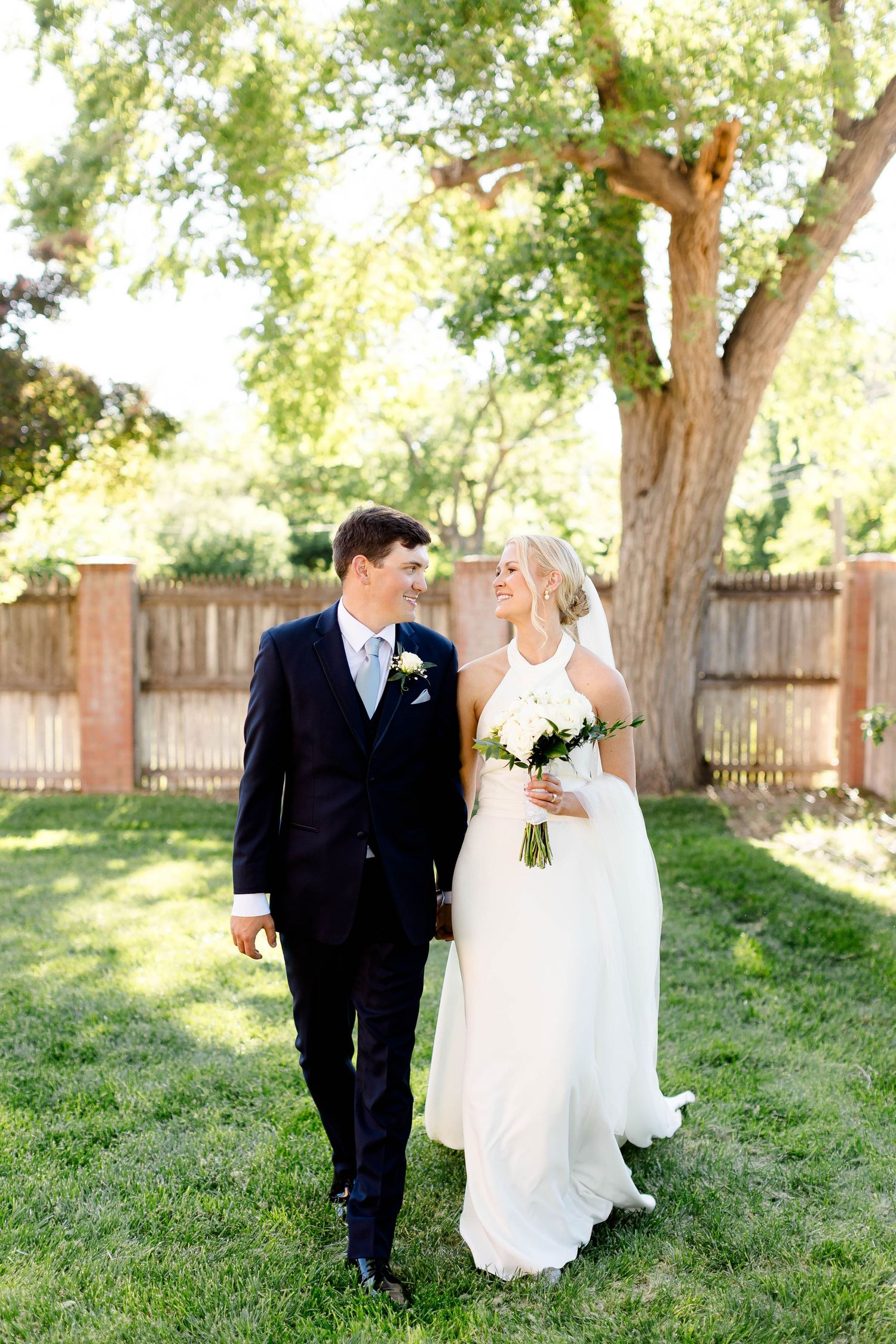 Newlyweds walking through a sunlit garden with trees in the background.