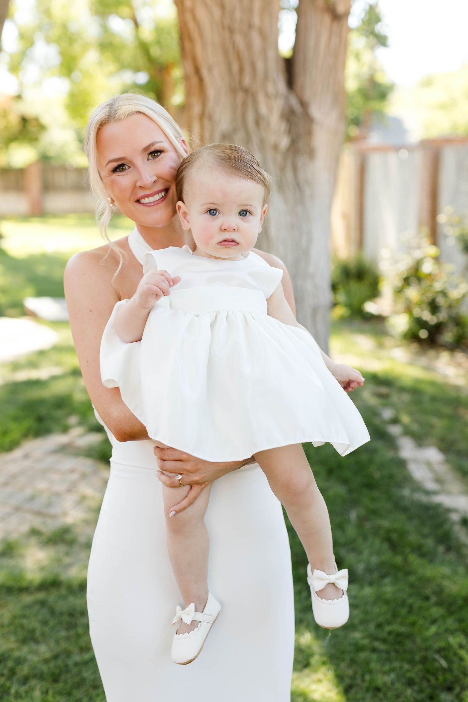 Baby in white dress being held against outdoor garden background.