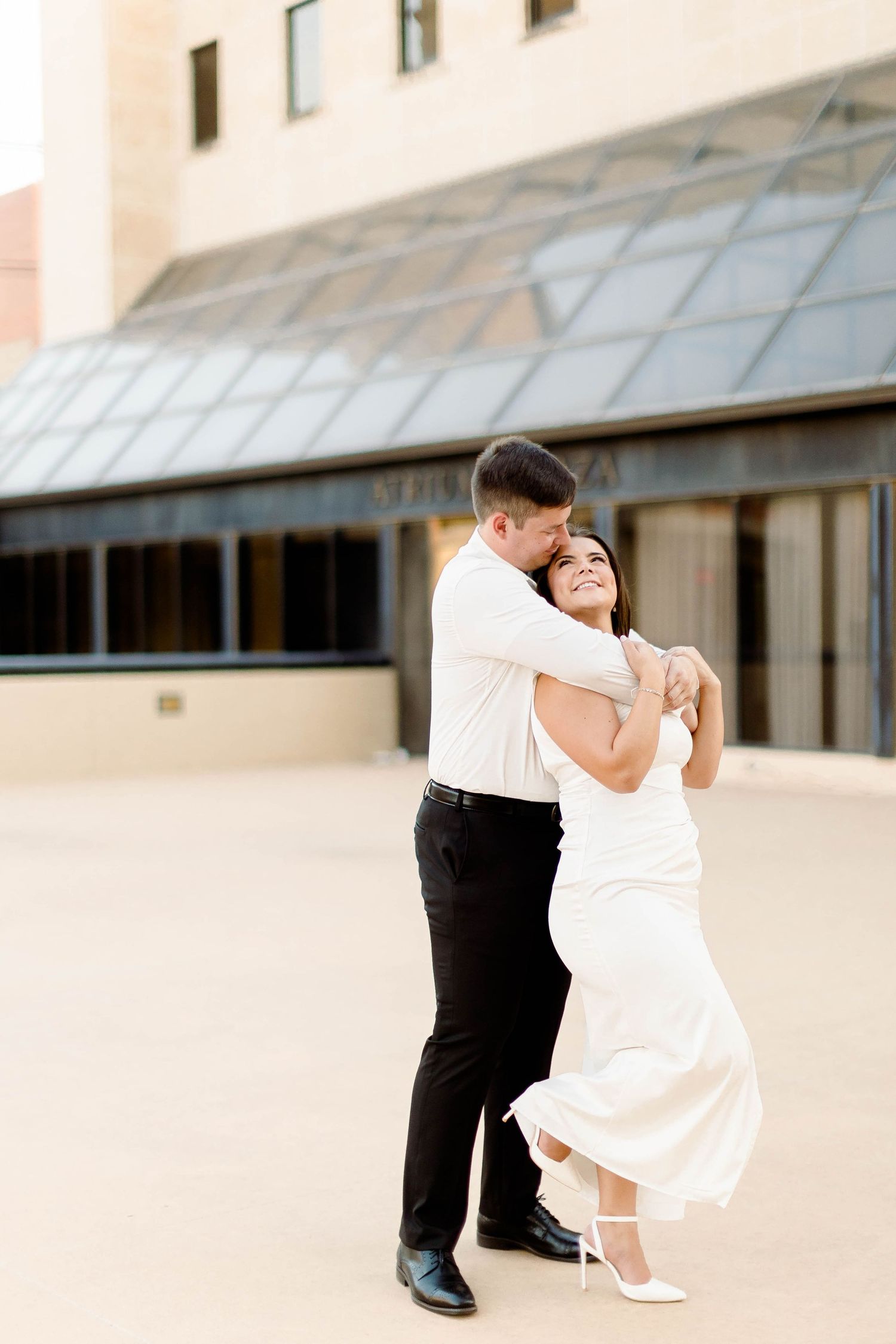 A couple in formal attire dance playfully against a modern building backdrop.
