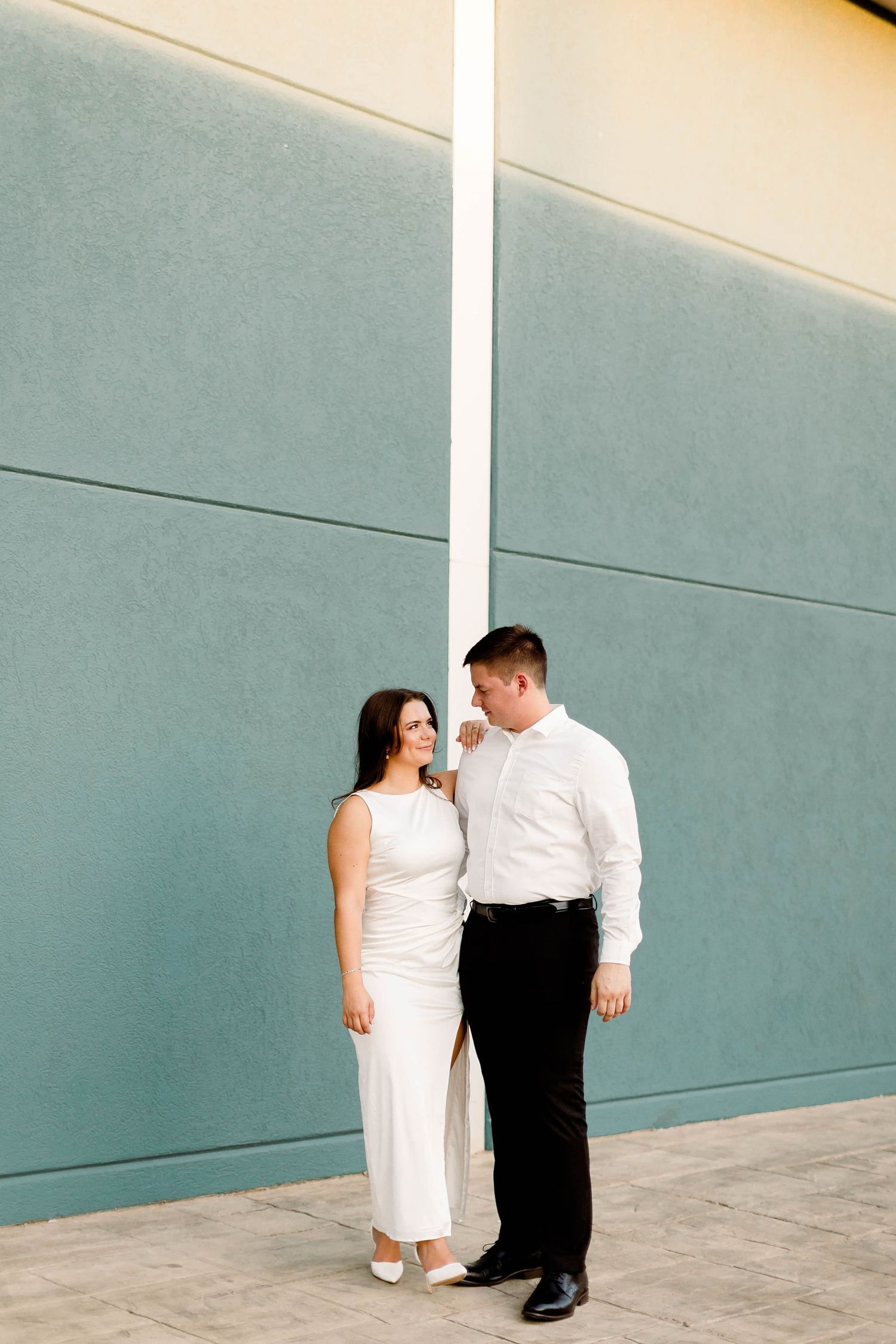 A couple in formal wear poses against a minimalist teal and cream wall.