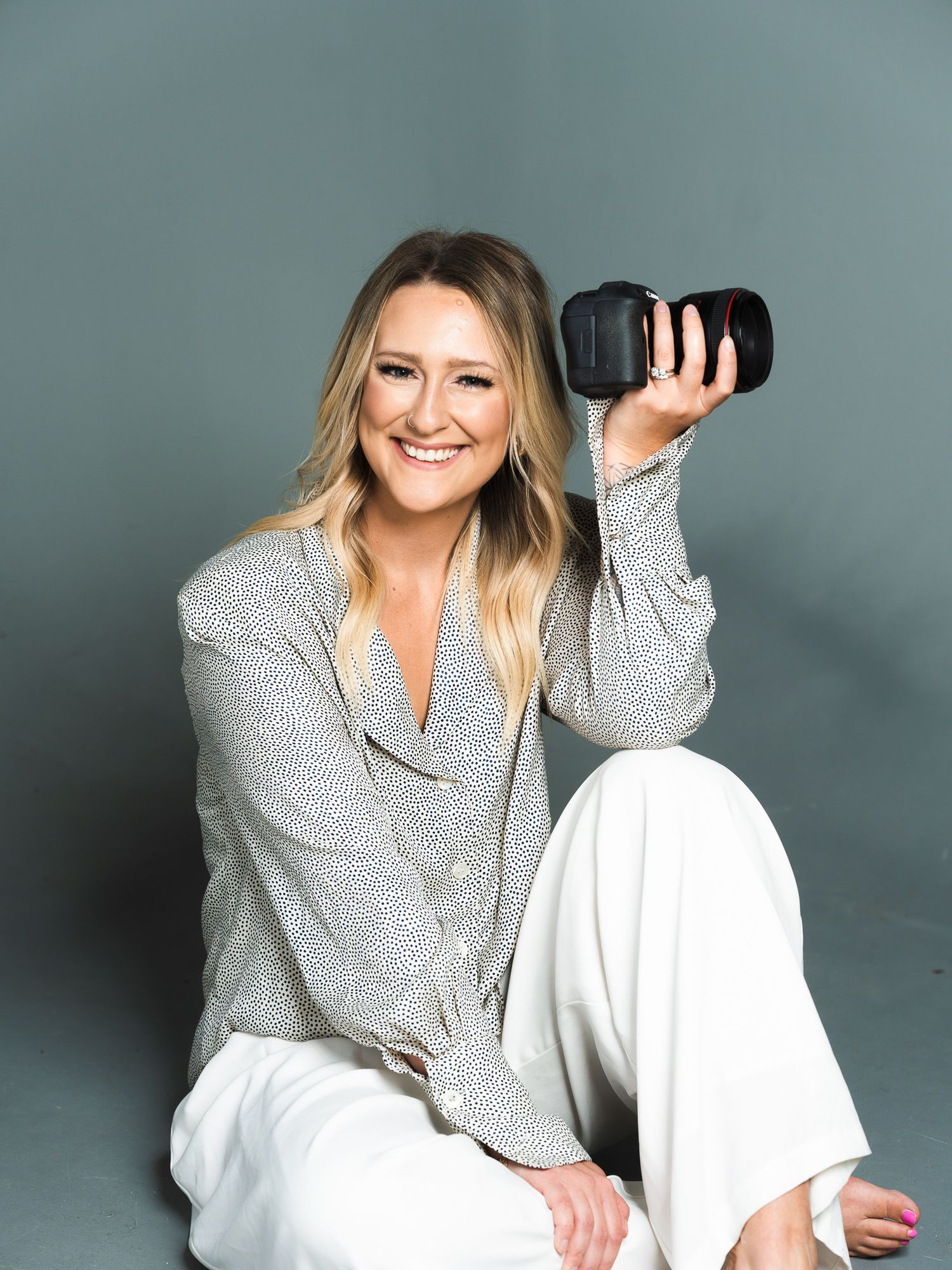 Professional photographer holding camera while posing in casual grey attire against studio backdrop.