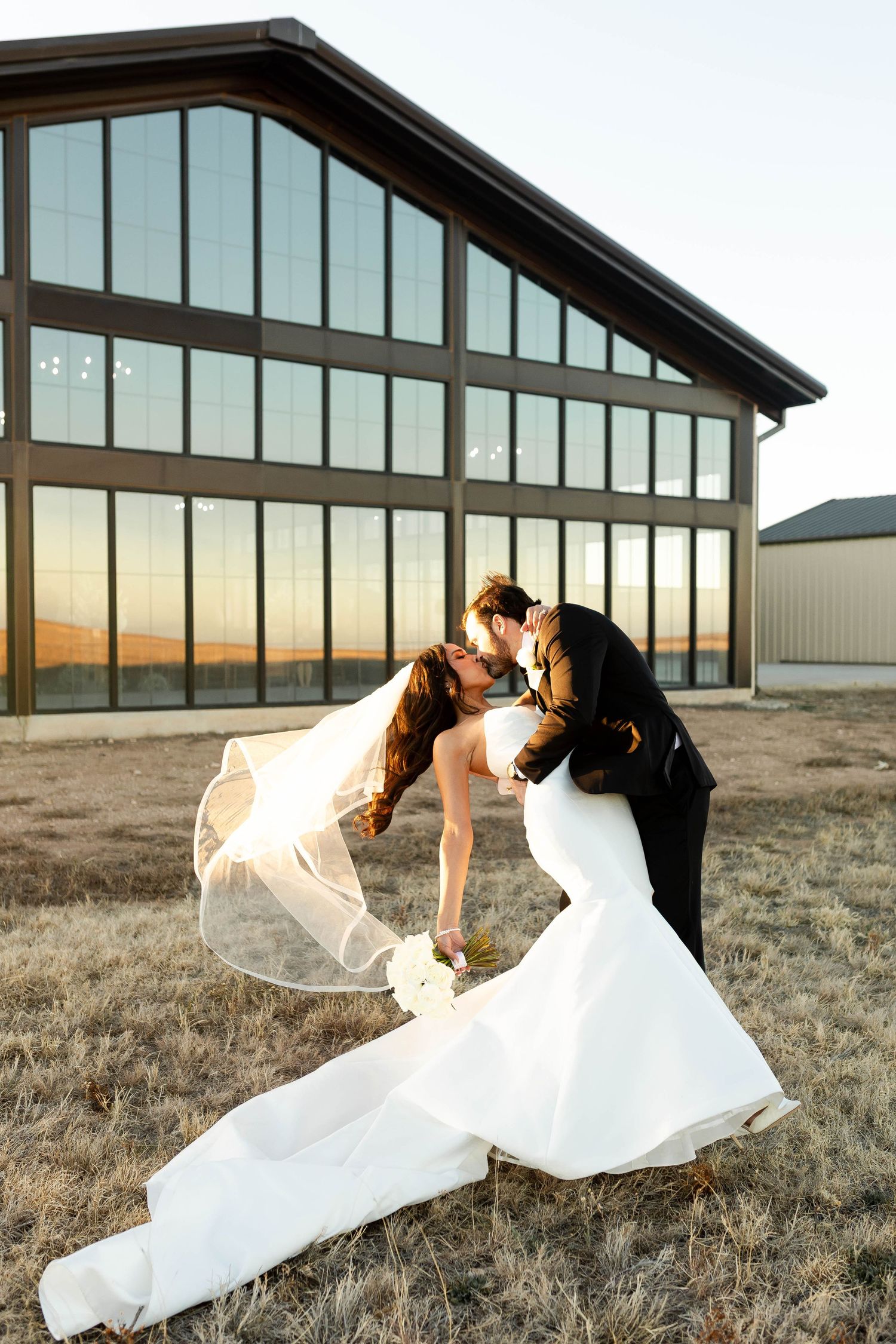 A romantic dip kiss between newlyweds outside a modern glass building at sunset.