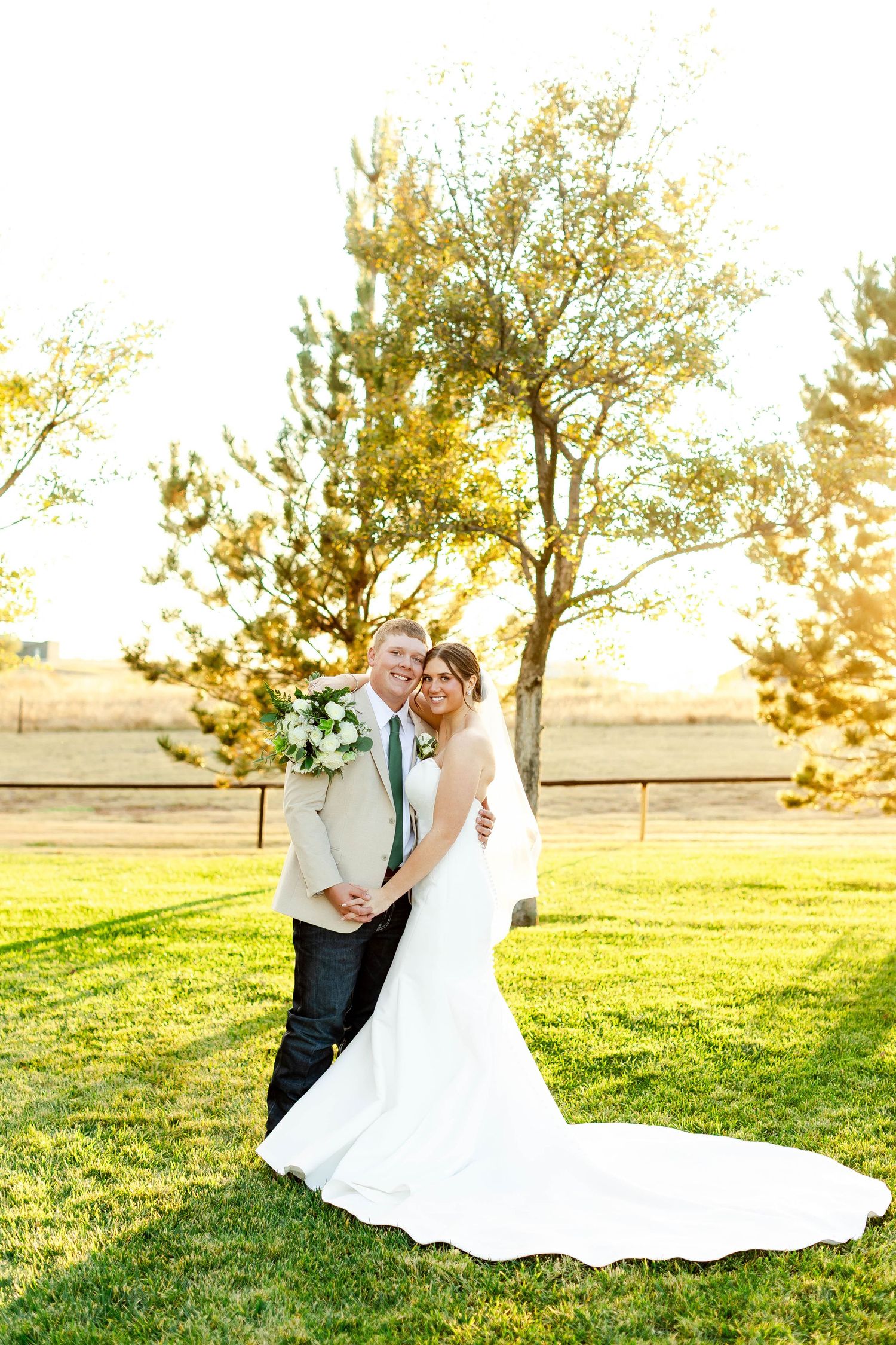 A wedding couple poses on grass at sunset with golden light filtering through trees.
