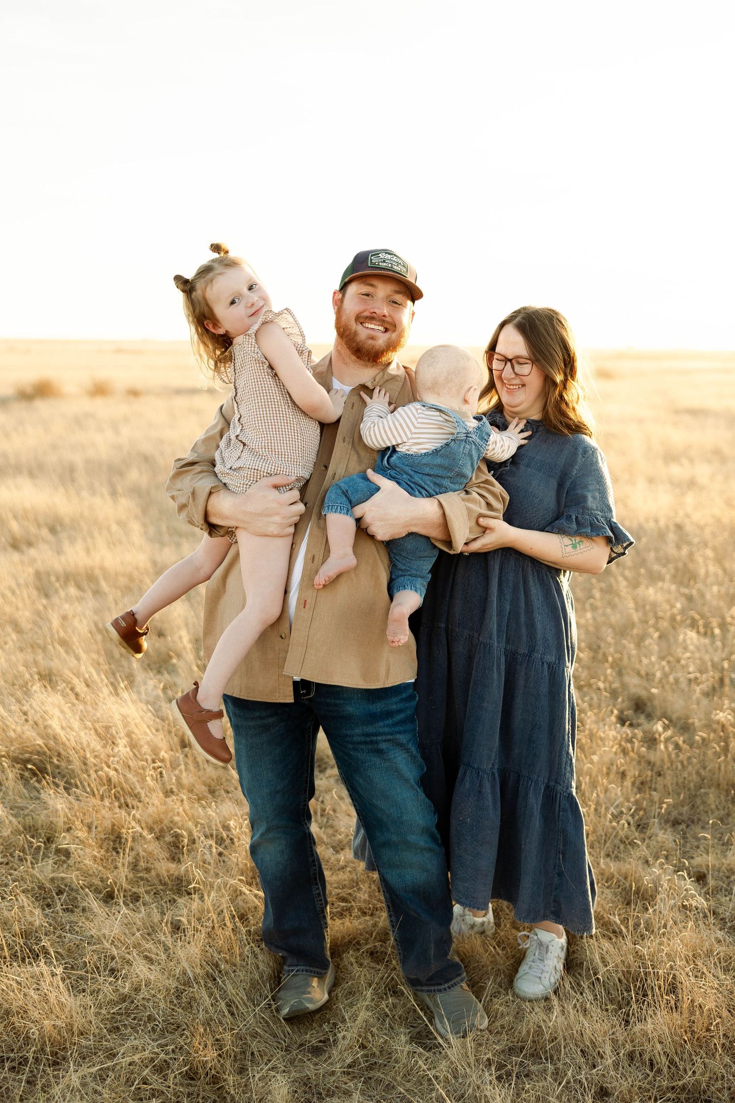 A family stands together in a golden field at sunset wearing casual clothing and embracing each other warmly.