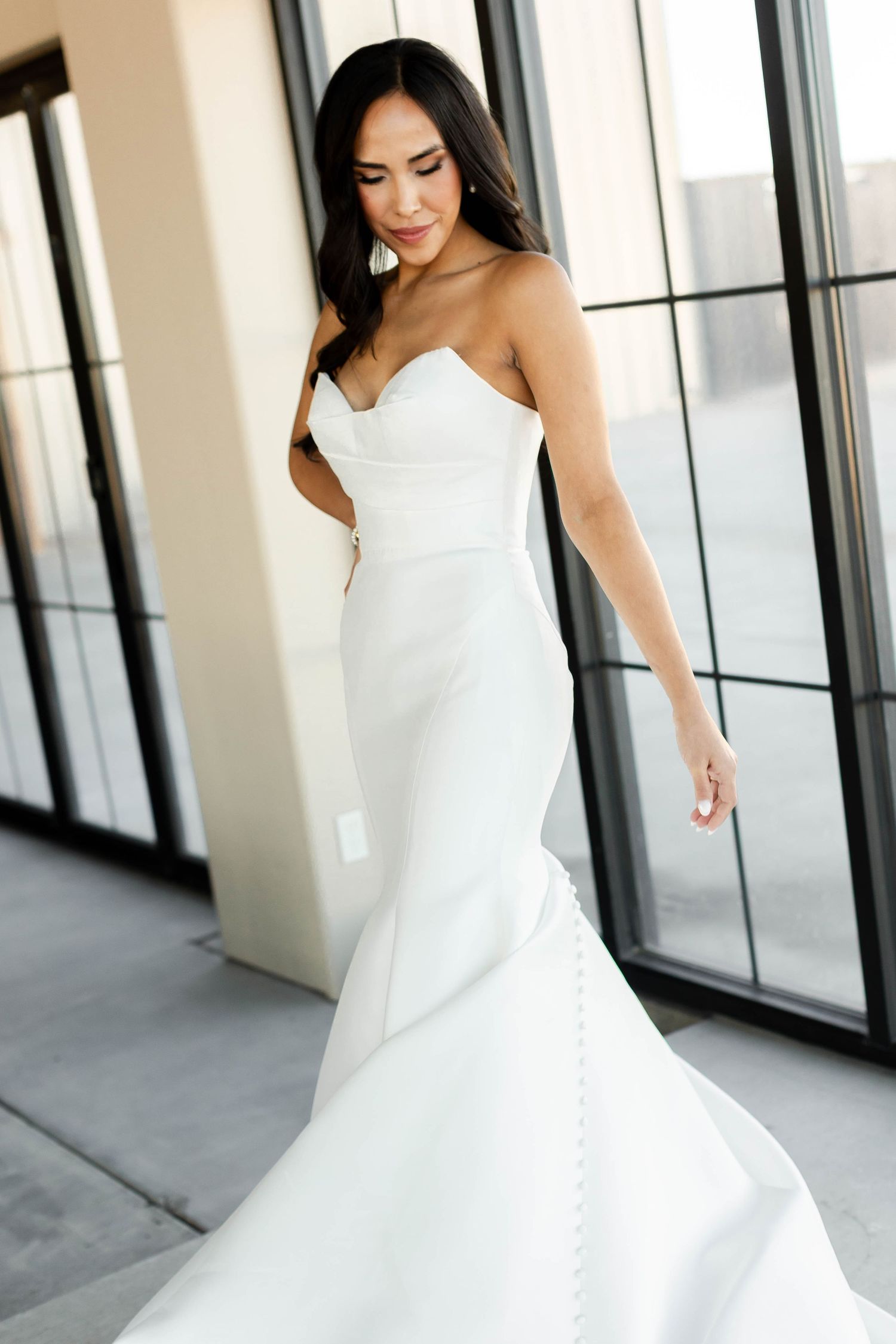 A bride poses in an elegant white fitted mermaid wedding gown against modern windows.