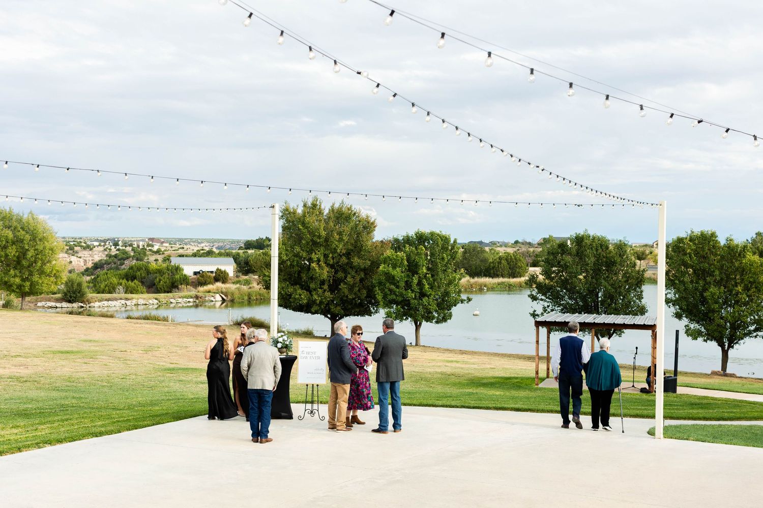 Wedding guests gather on a paved path near a lake with string lights overhead and trees in the background.