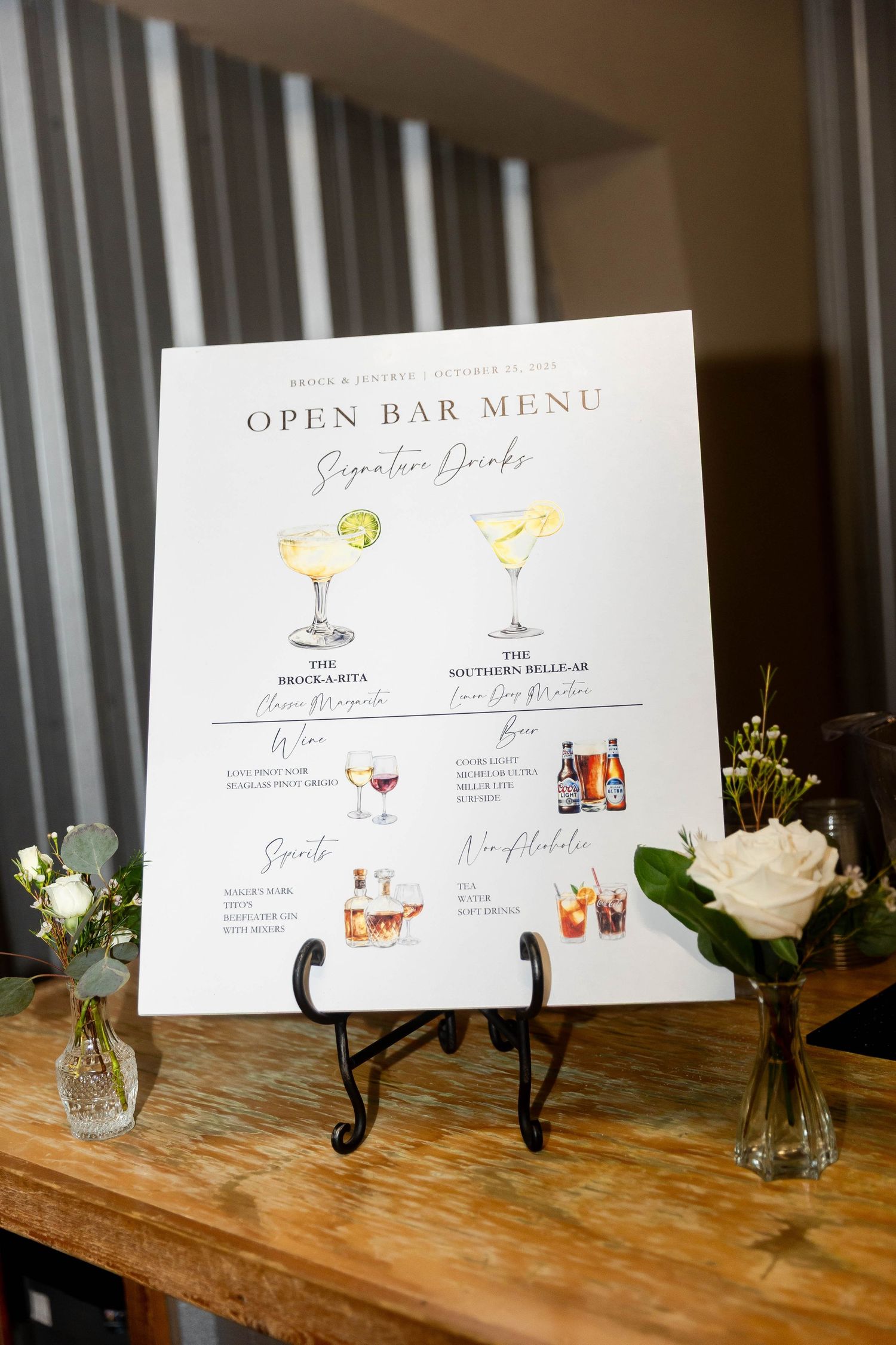 Wedding bar menu displayed on wooden stands with illustrated cocktail drawings and white floral accents.