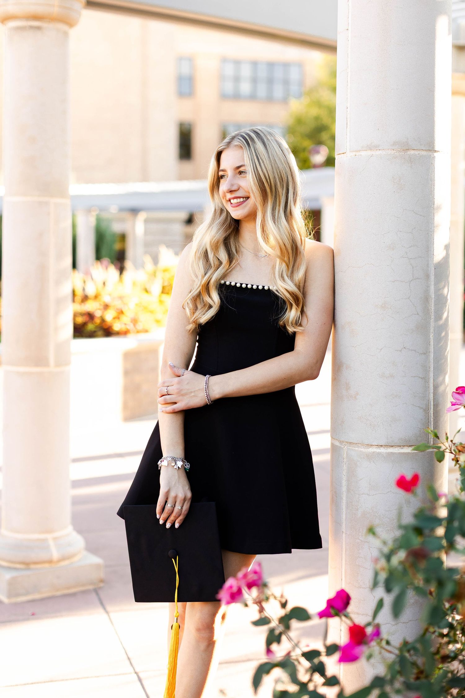 Person in black dress poses between white columns with pink flowers. WT senior