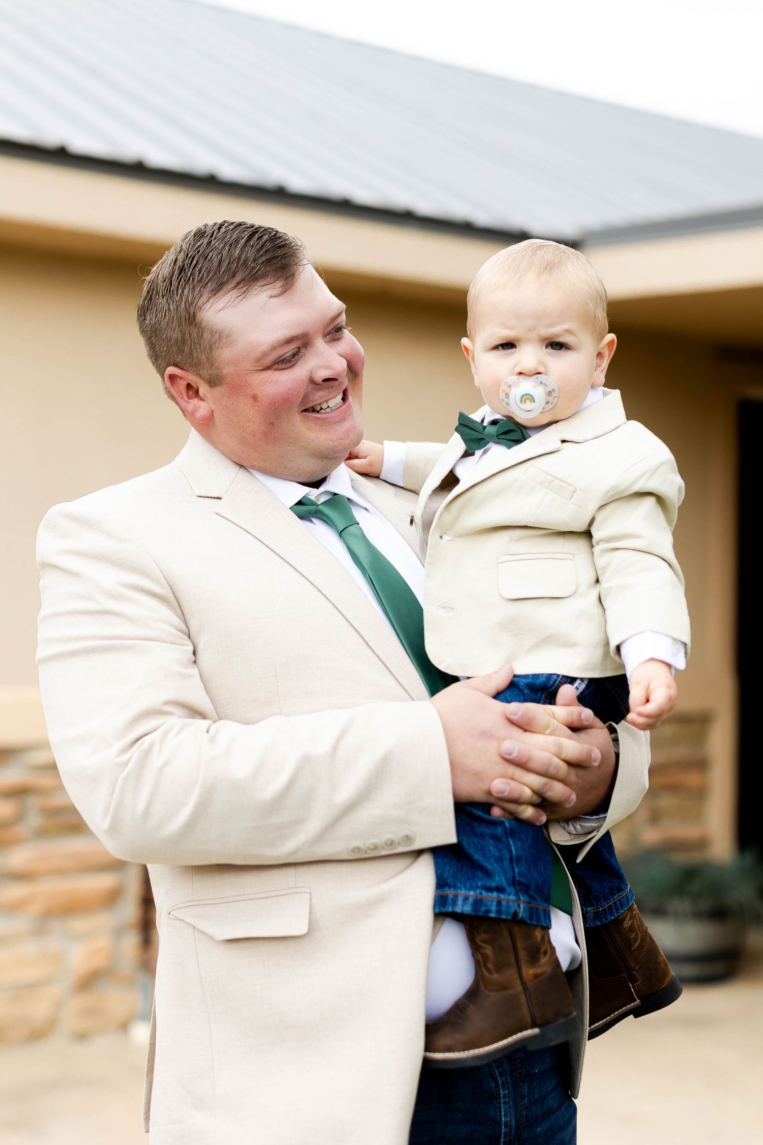 A small baby wearing jeans and a white shirt is held by someone in a cream colored suit jacket.