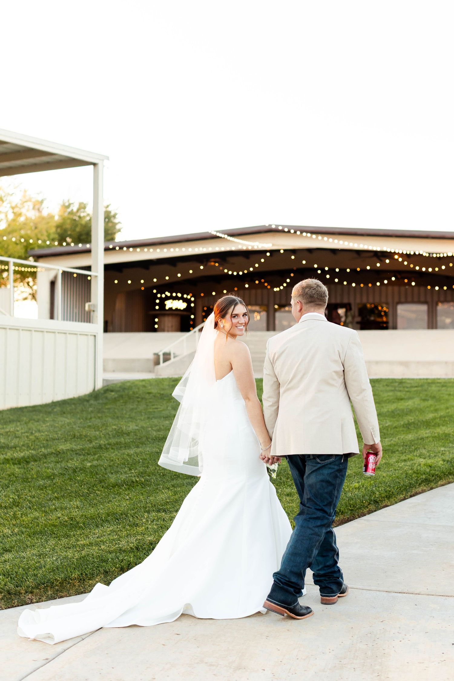 Newlyweds walk hand in hand along a concrete path outside their wedding venue at dusk.