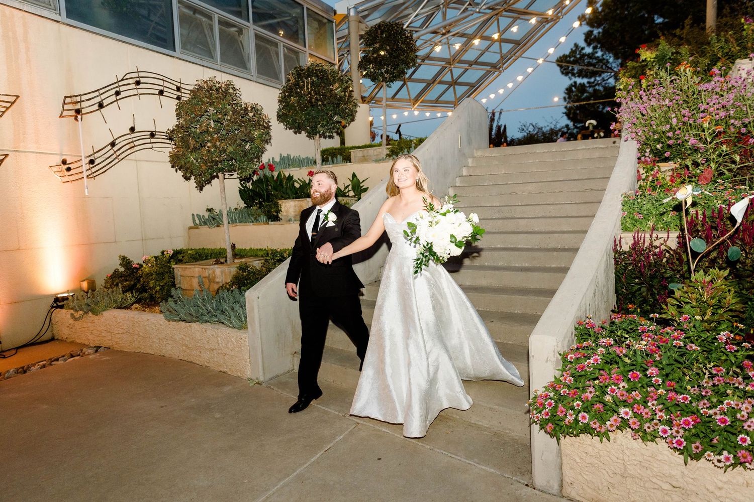 A couple dances together during their wedding celebration in a romantic garden setting with string lights overhead.