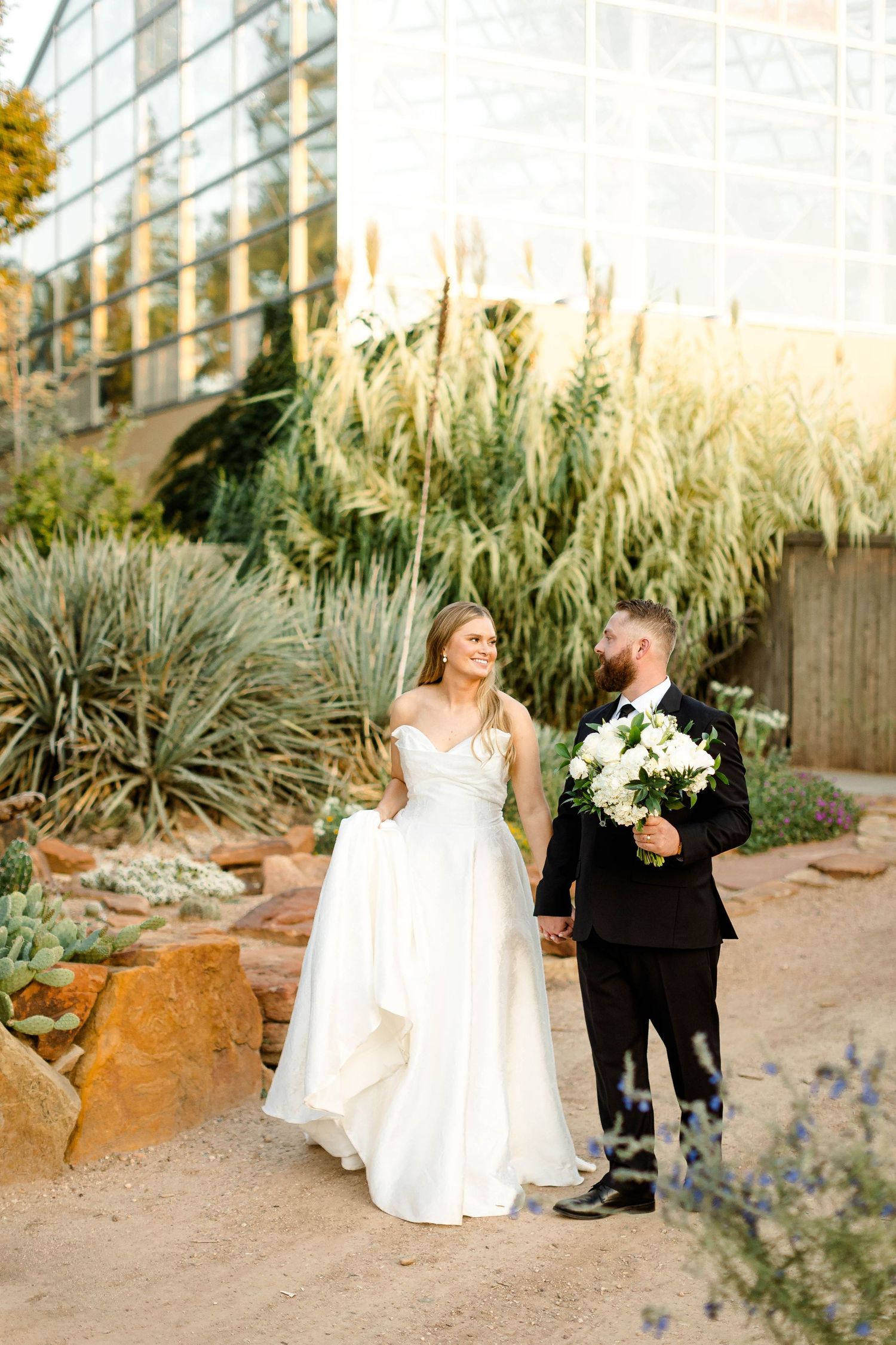 Newlyweds walking through garden path with ornamental grasses and modern architecture.