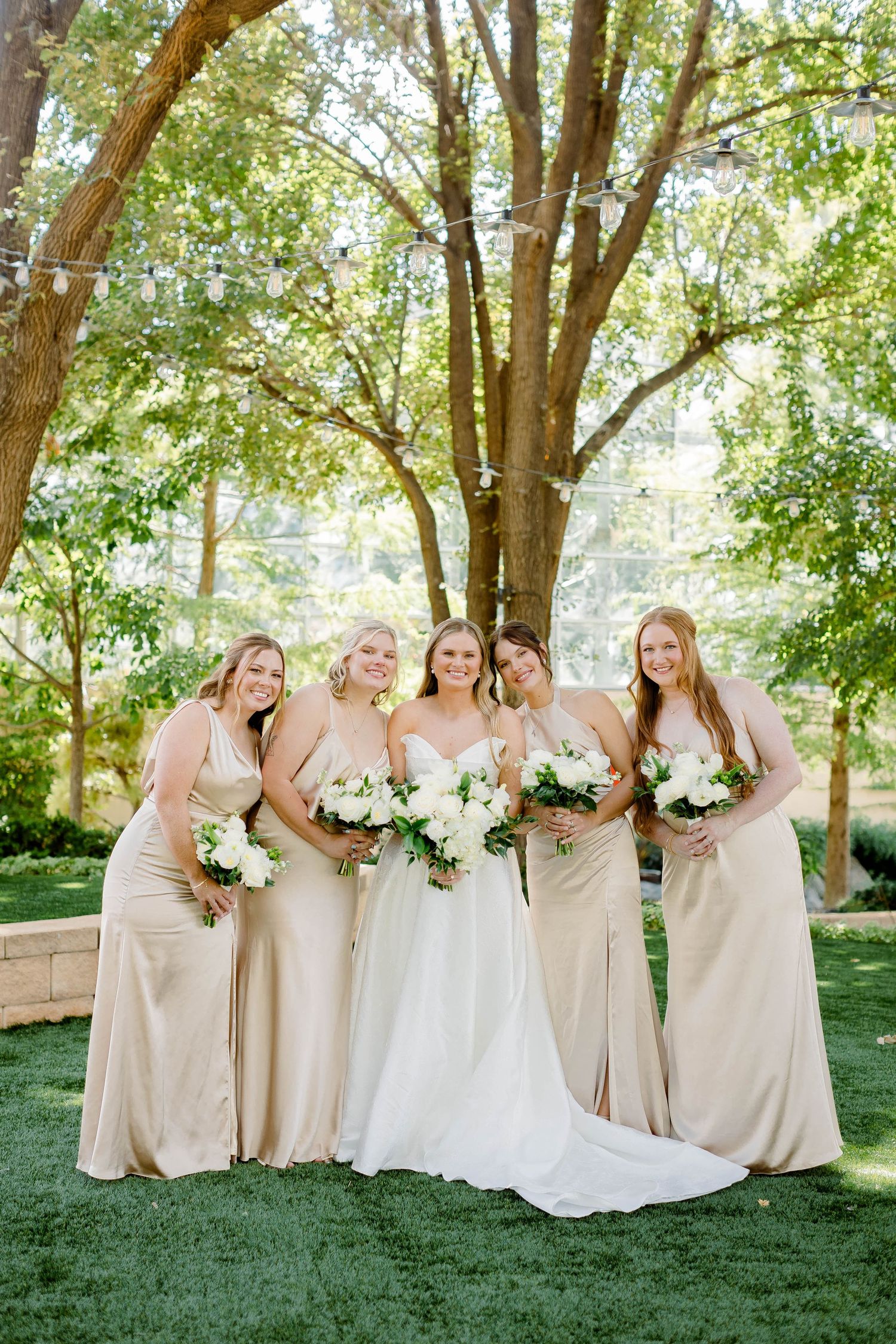 Bridal party in champagne-colored dresses pose with white bouquets under trees in an outdoor garden setting.