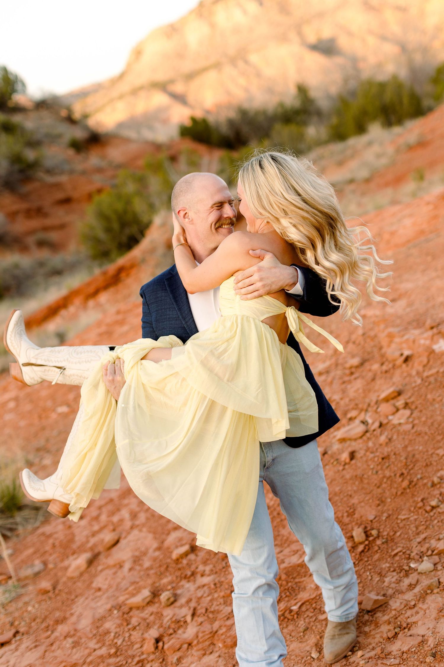 A couple dances playfully against a desert backdrop with red rocks and mountains at sunset.